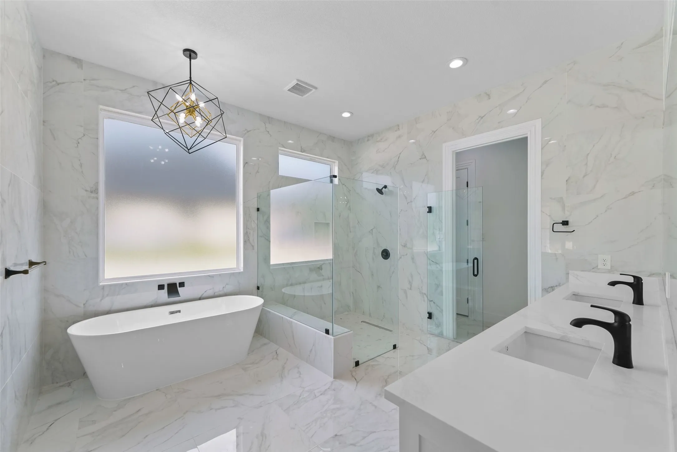 Freestanding soaking tub, frameless glass shower enclosure, dual vanity with undermount sinks, geometric chandelier, and large format wall and floor tiling