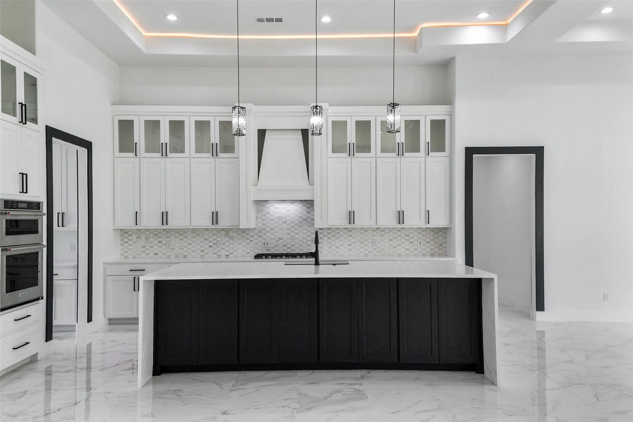 Gourmet kitchen featuring a waterfall island with a light countertop, white upper cabinetry with glass inserts, stainless steel double ovens, and a tile backsplash