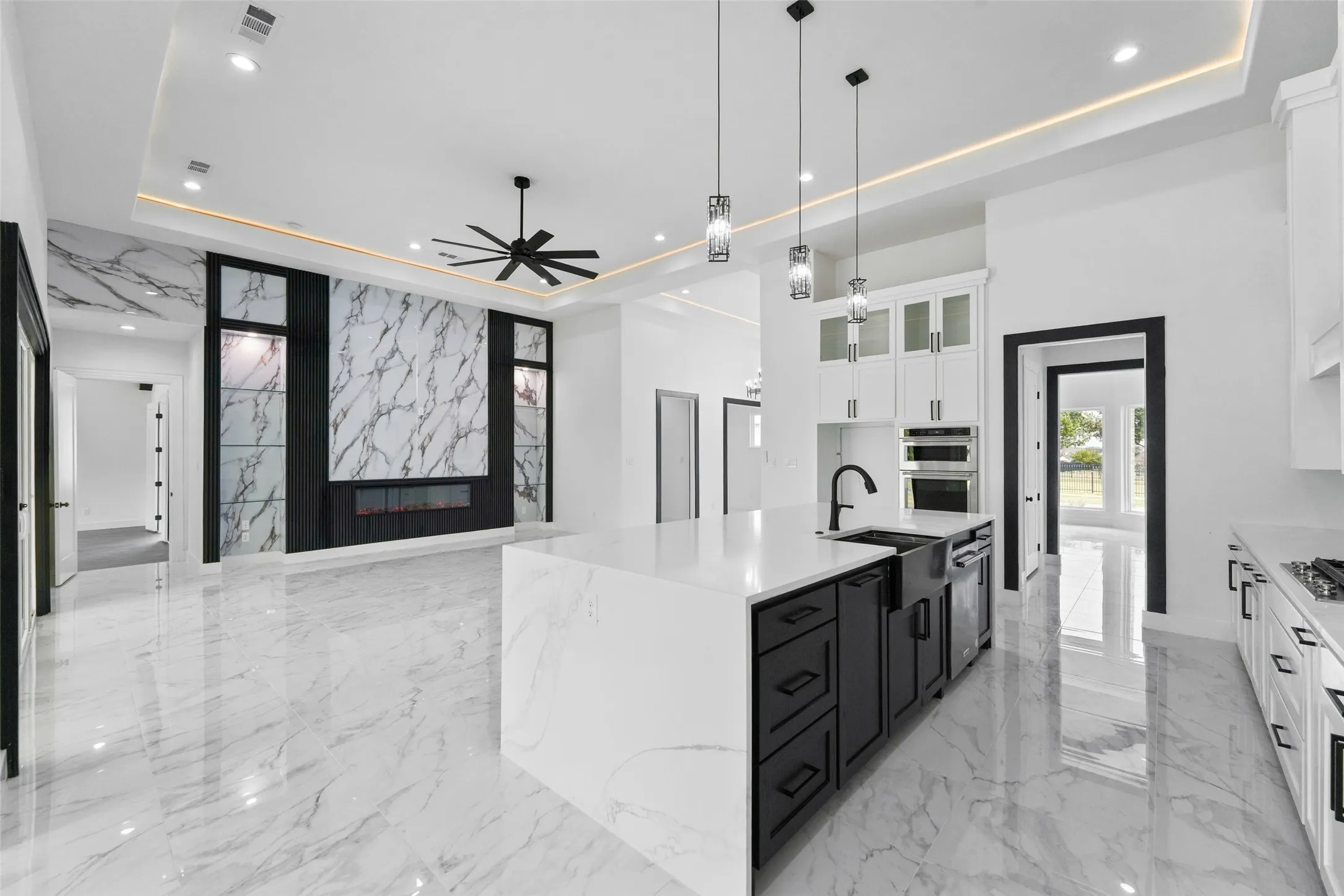 Spacious open-concept interior with large-format marble-look flooring, a kitchen island featuring a black farmhouse sink and pendant lighting, and a modern fireplace with a marble-look surround