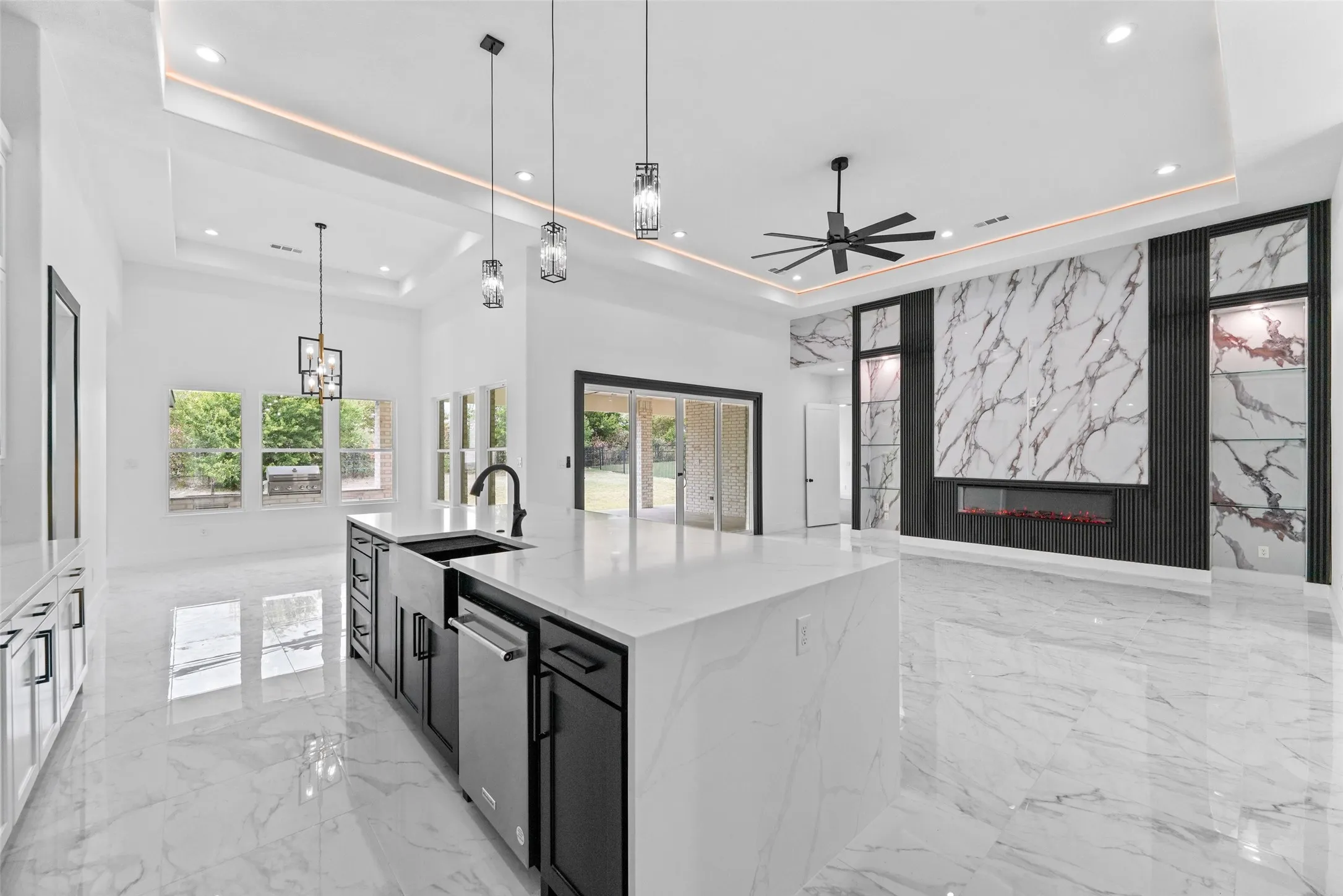 Spacious kitchen and living area featuring a large central island with an integrated sink and dishwasher, white marble-look flooring, recessed and pendant lighting, a tray ceiling, and a prominent fireplace with a decorative stone-look surround