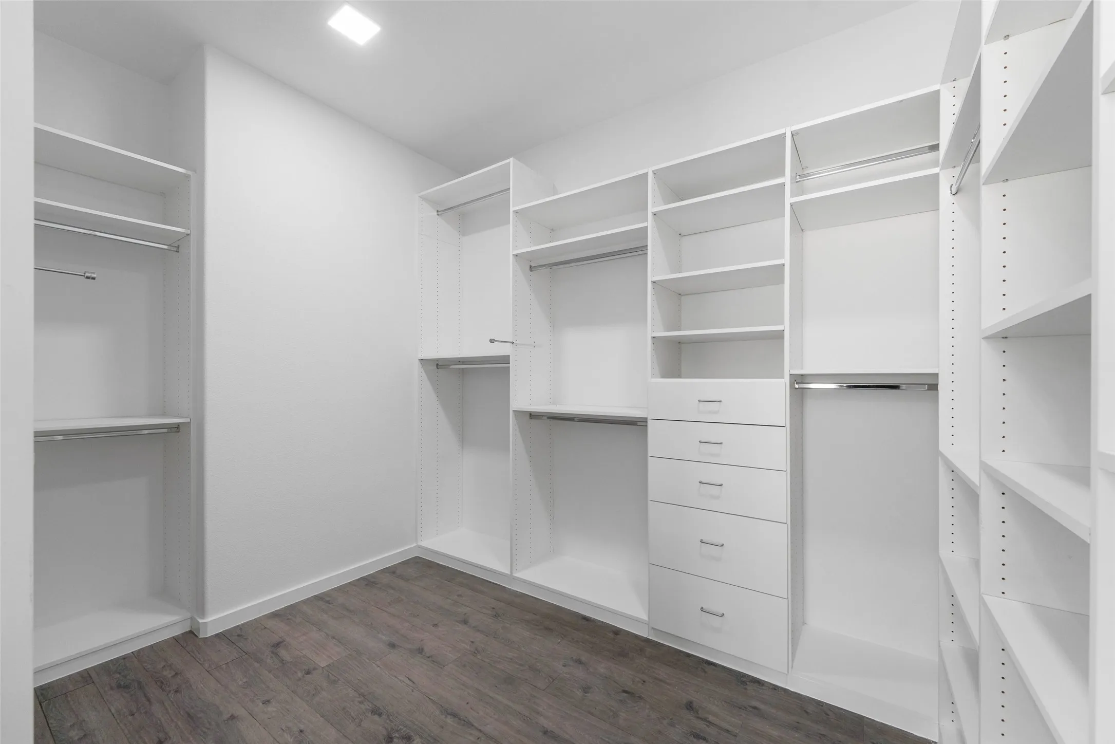 Spacious walk-in closet featuring custom white cabinetry with multiple drawers, open shelving throughout, hanging rods at varying heights, and wood-finish flooring