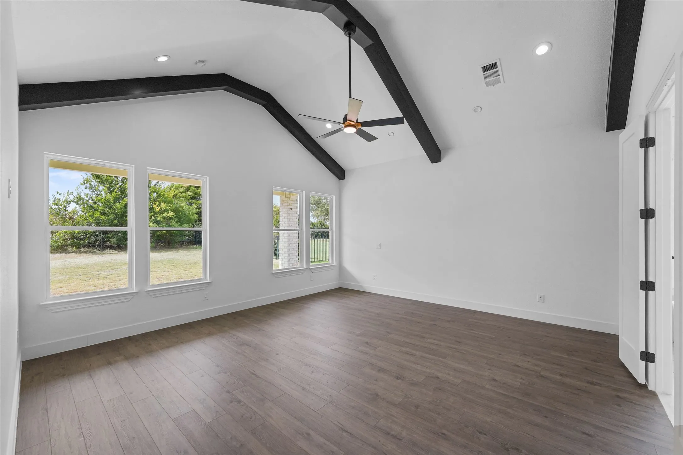 Bright, open room with cathedral ceiling and exposed dark wood beams, featuring three large windows with natural light and garden views
