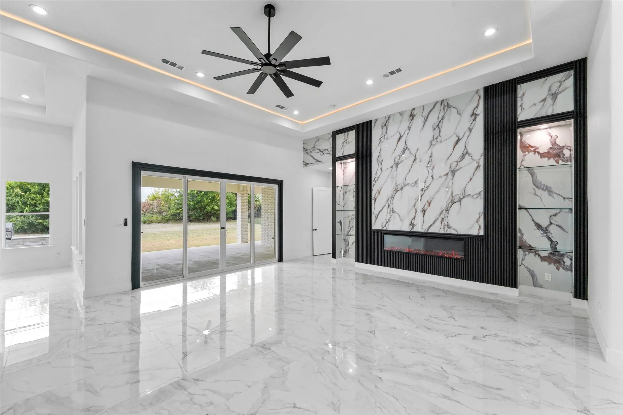 Polished marble-look tile flooring, electric fireplace with stone-look surround, tray ceiling with cove lighting, and sliding glass doors to the exterior