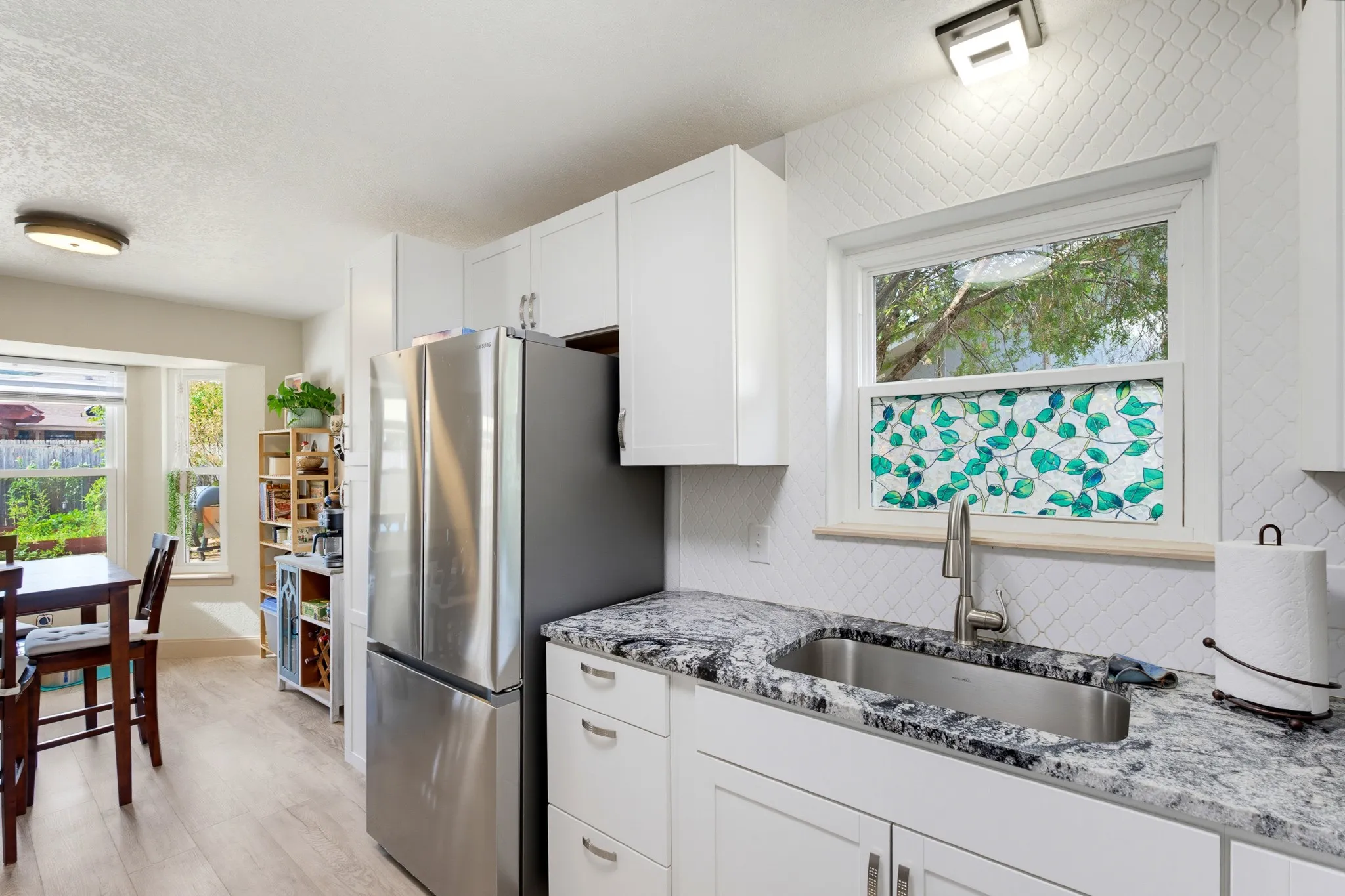 White cabinetry with stainless steel hardware, stainless steel refrigerator, undermount sink with a gooseneck faucet, light-toned wood-finish flooring, and a patterned backsplash