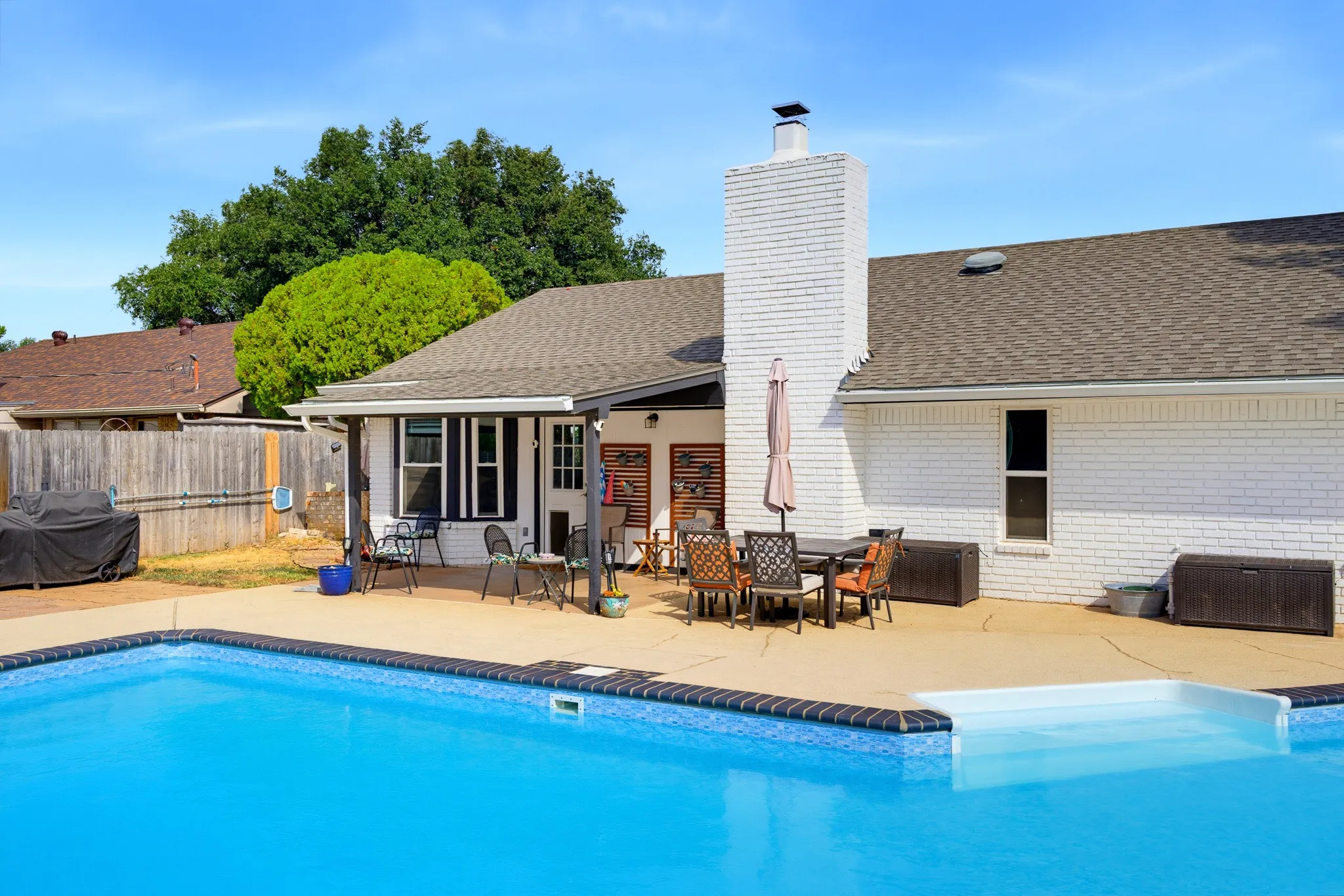 Backyard featuring an in-ground swimming pool, expansive concrete patio, and a white brick-clad exterior with a prominent chimney