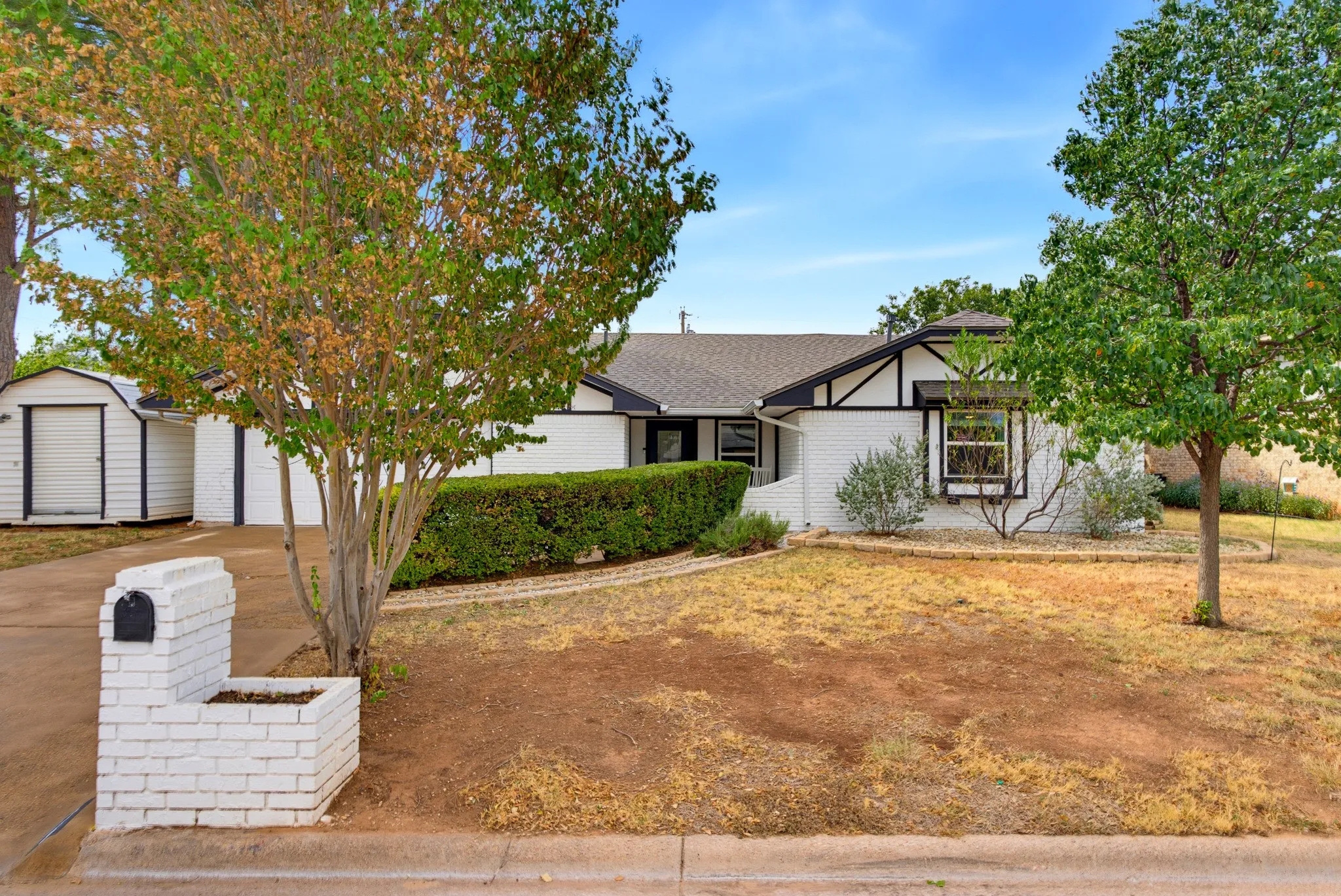 White brick exterior with black trim detailing, mature trees, a driveway, and a detached storage shed