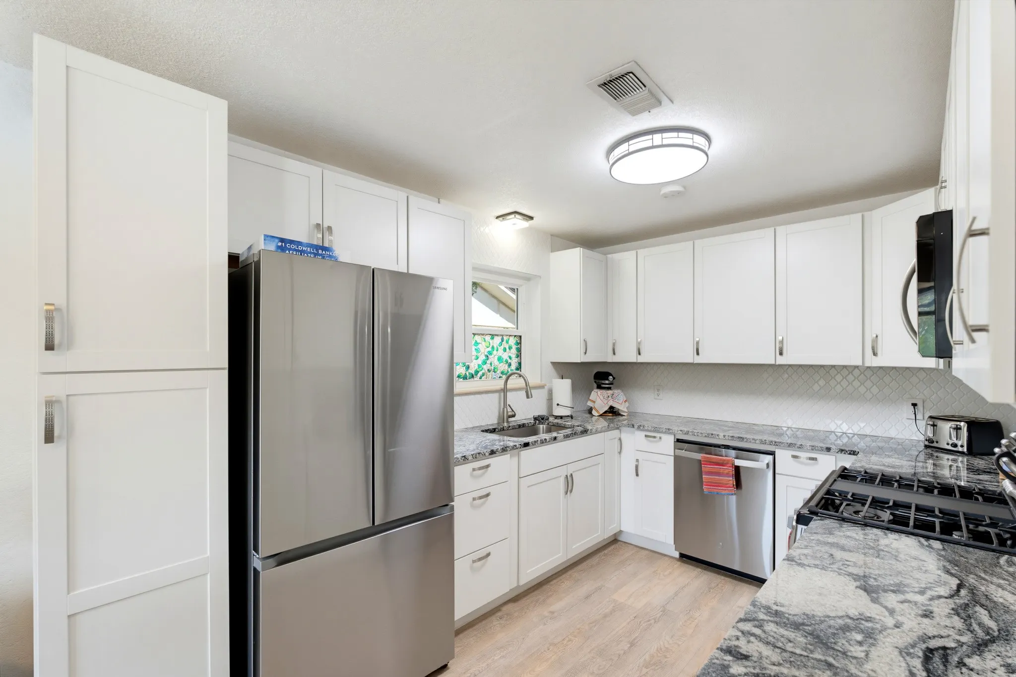 White shaker-style cabinetry, stainless steel appliances, patterned backsplash, light wood-finish flooring, and a stone countertop