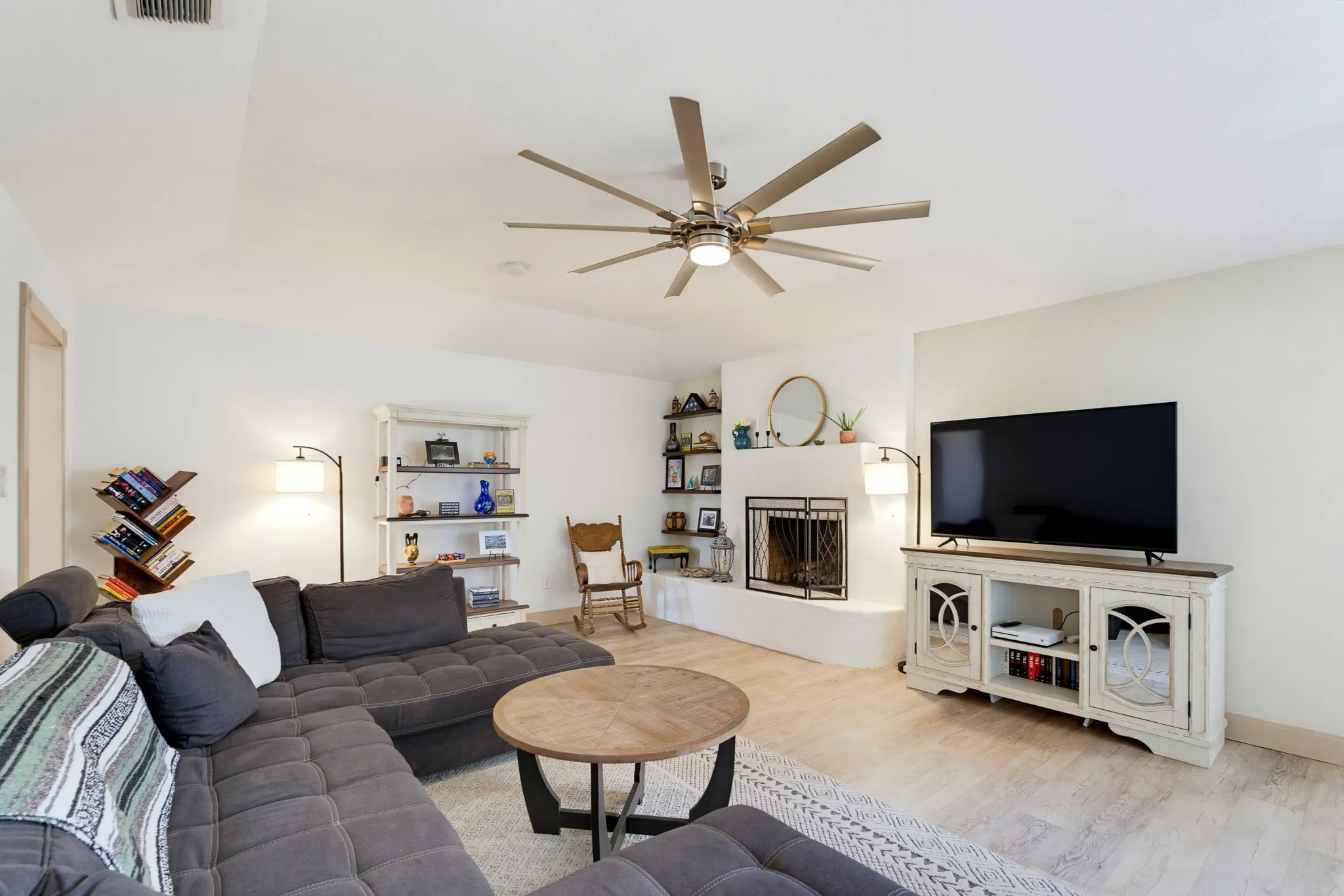 Corner fireplace with metal grate, light wood-finish flooring, white walls, and a ceiling fan with integrated lighting