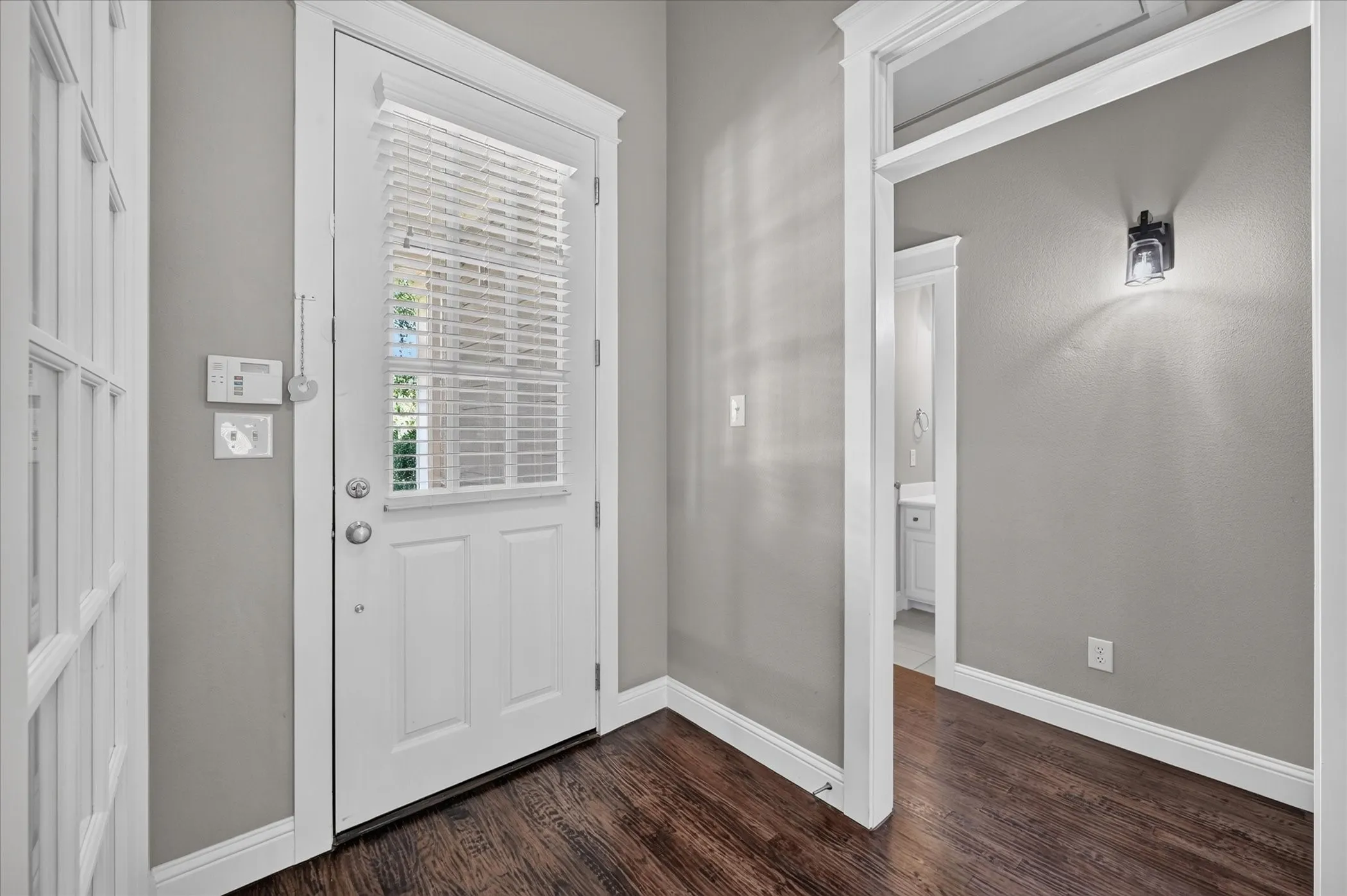Rich wood-finish flooring with crisp white trim, a transom window door with privacy blinds, and a built-in wall sconce