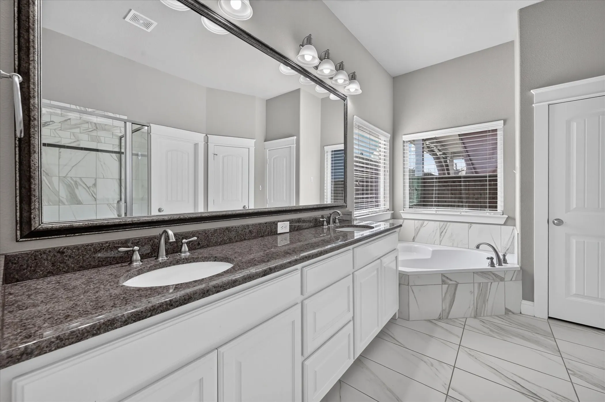 Dual vanity with undermount sinks and dark stone countertops, white cabinetry, large framed mirror, built-in tub with tile surround, and large format tile flooring