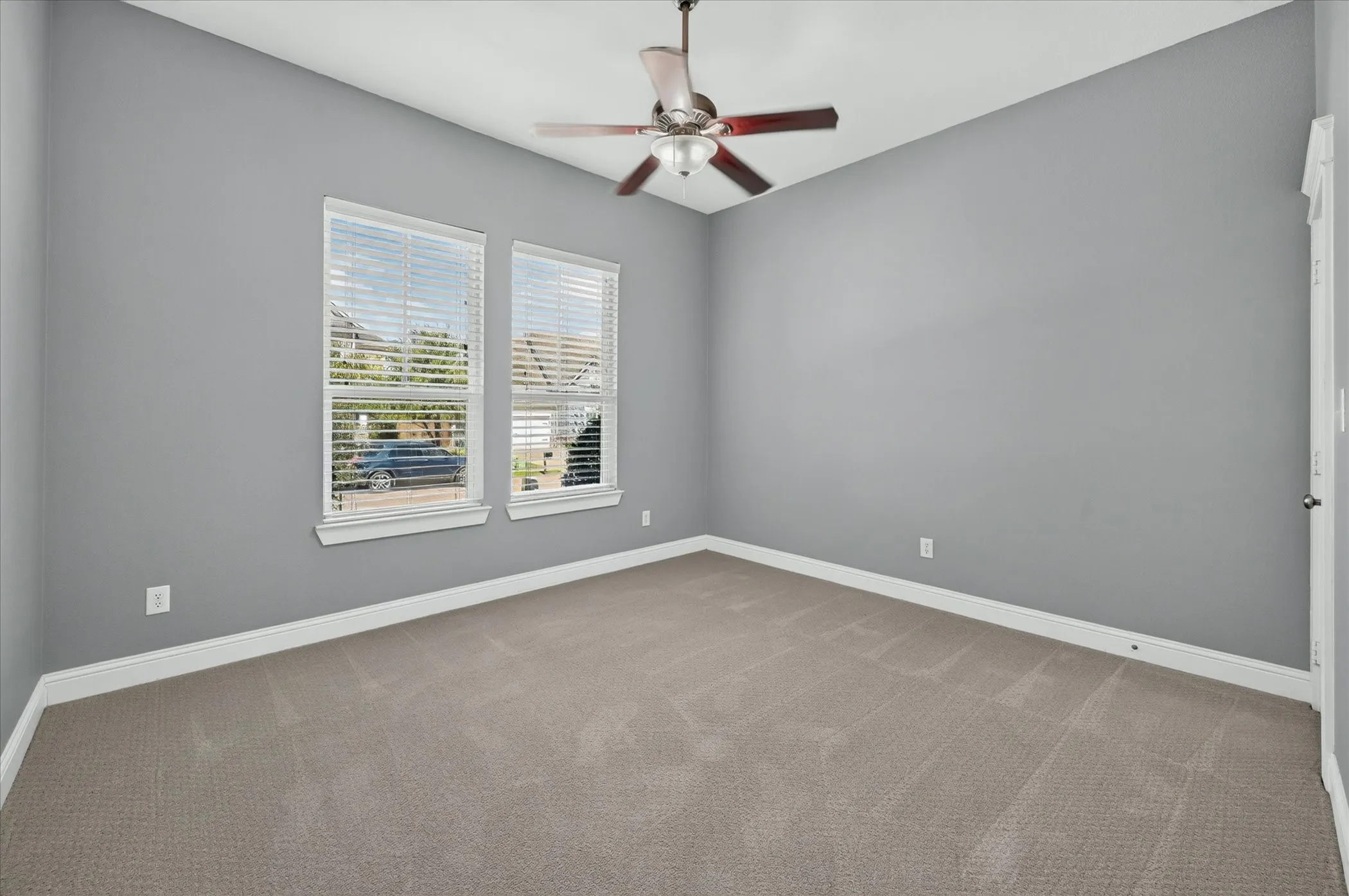 Dual windows with horizontal blinds, overhead ceiling fan with light fixture, gray wall paint, and neutral tone carpet flooring