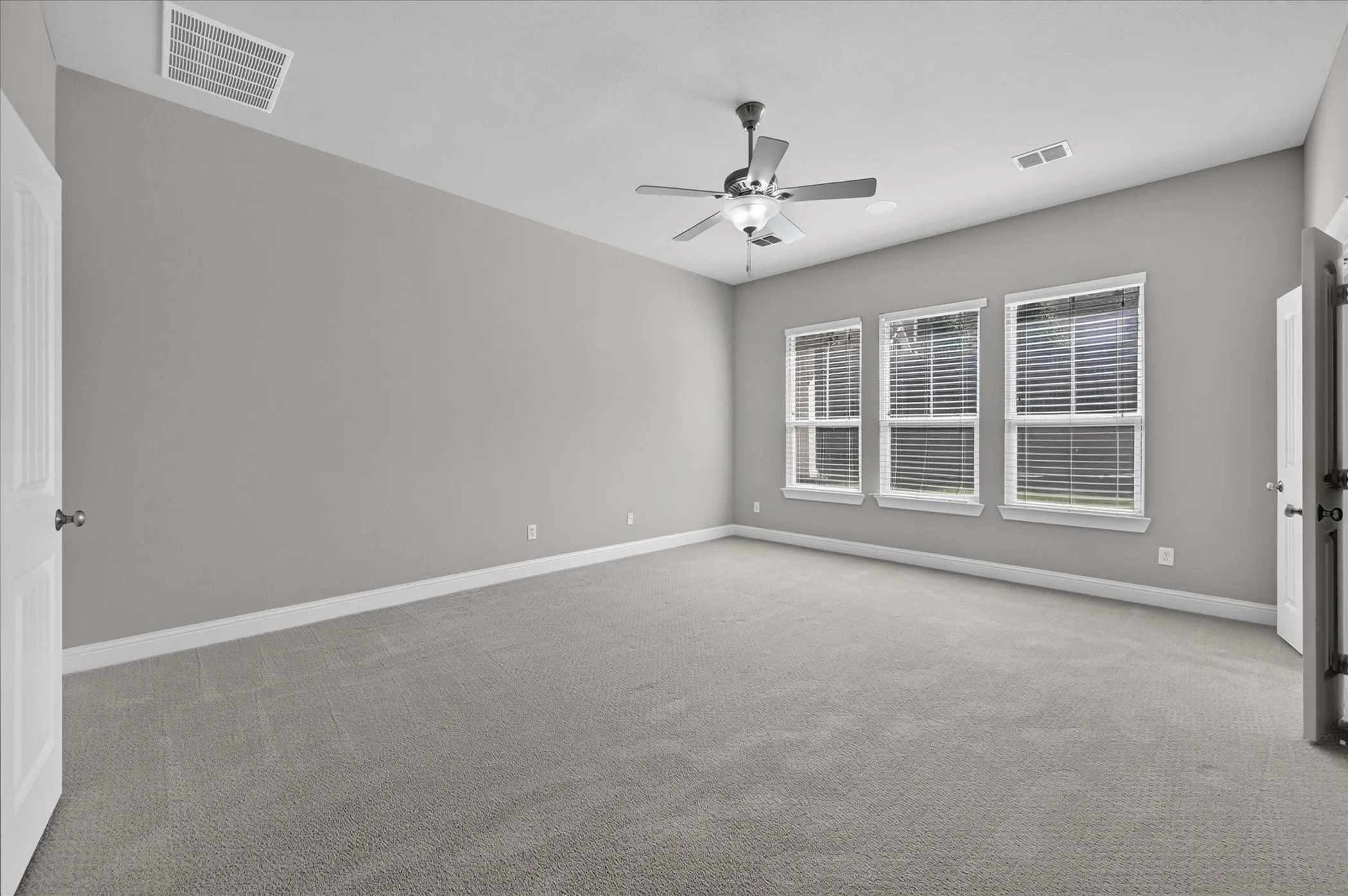 Spacious room featuring gray carpet, three large windows with blinds, a ceiling fan, and white trim
