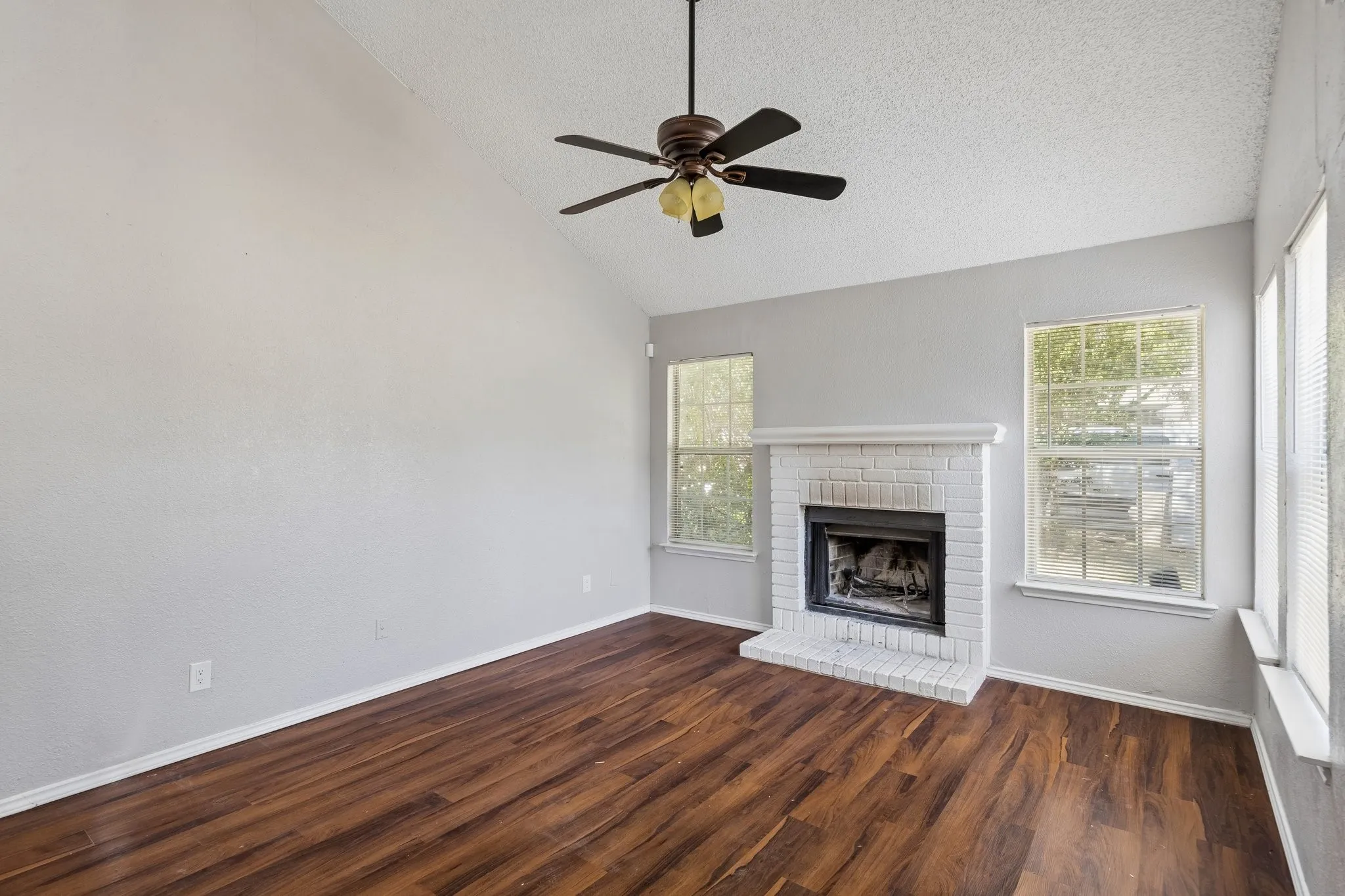 Vaulted ceiling with a ceiling fan, wood-finish flooring, white painted brick fireplace with hearth, and multiple windows
