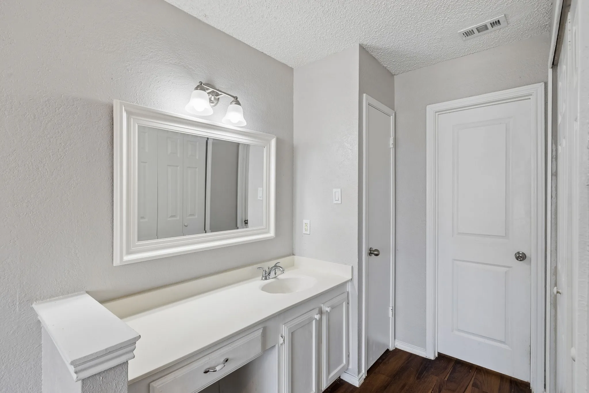 White vanity with integrated sink, framed mirror, two-light vanity fixture, and dark wood-finish​​‌​​​​‌​​‌‌​‌‌​​​‌‌​‌​‌​‌​​​‌​​ flooring