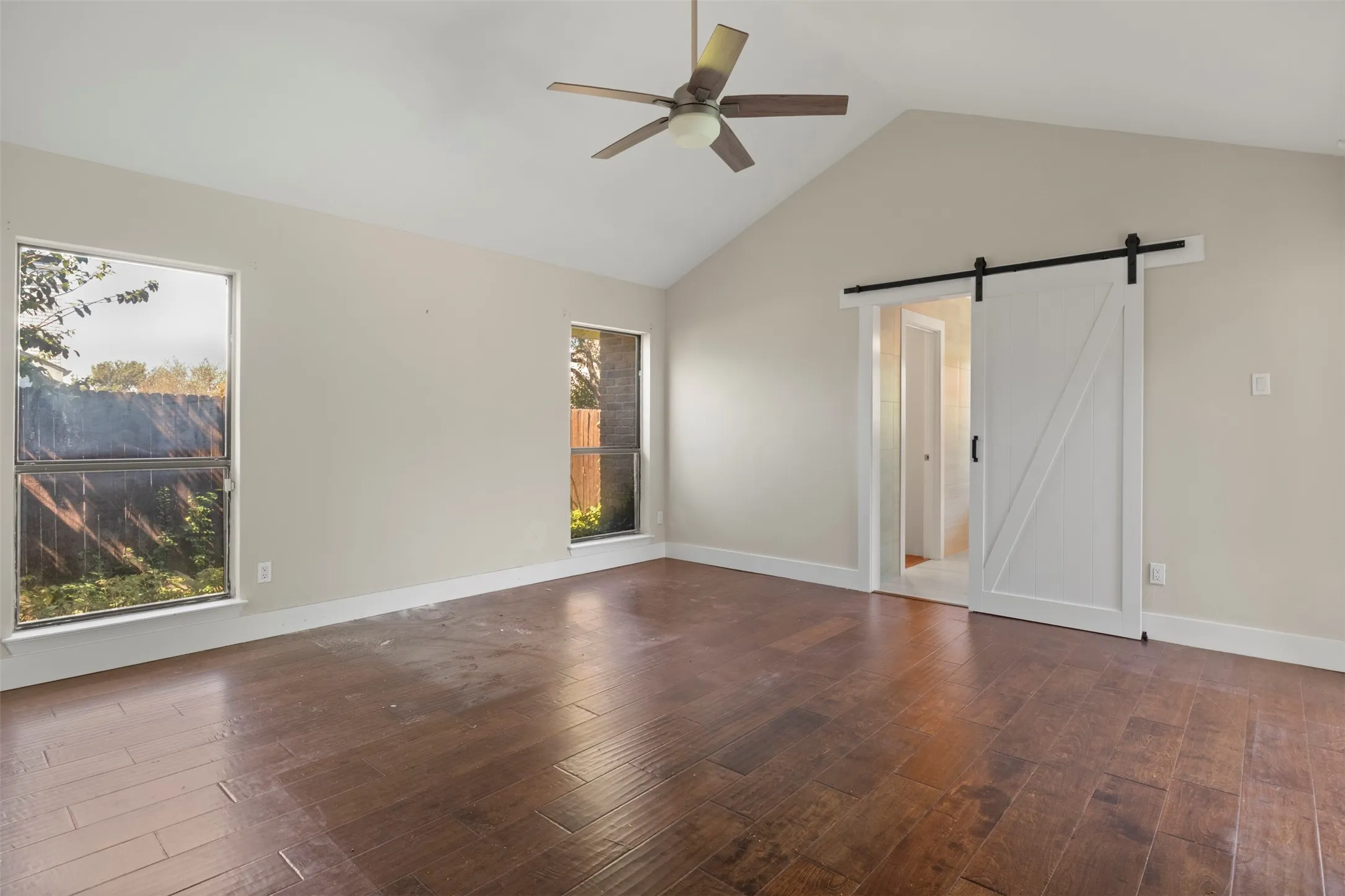 Vaulted ceiling with a ceiling fan, wood-finish flooring, two large windows, and a white barn door