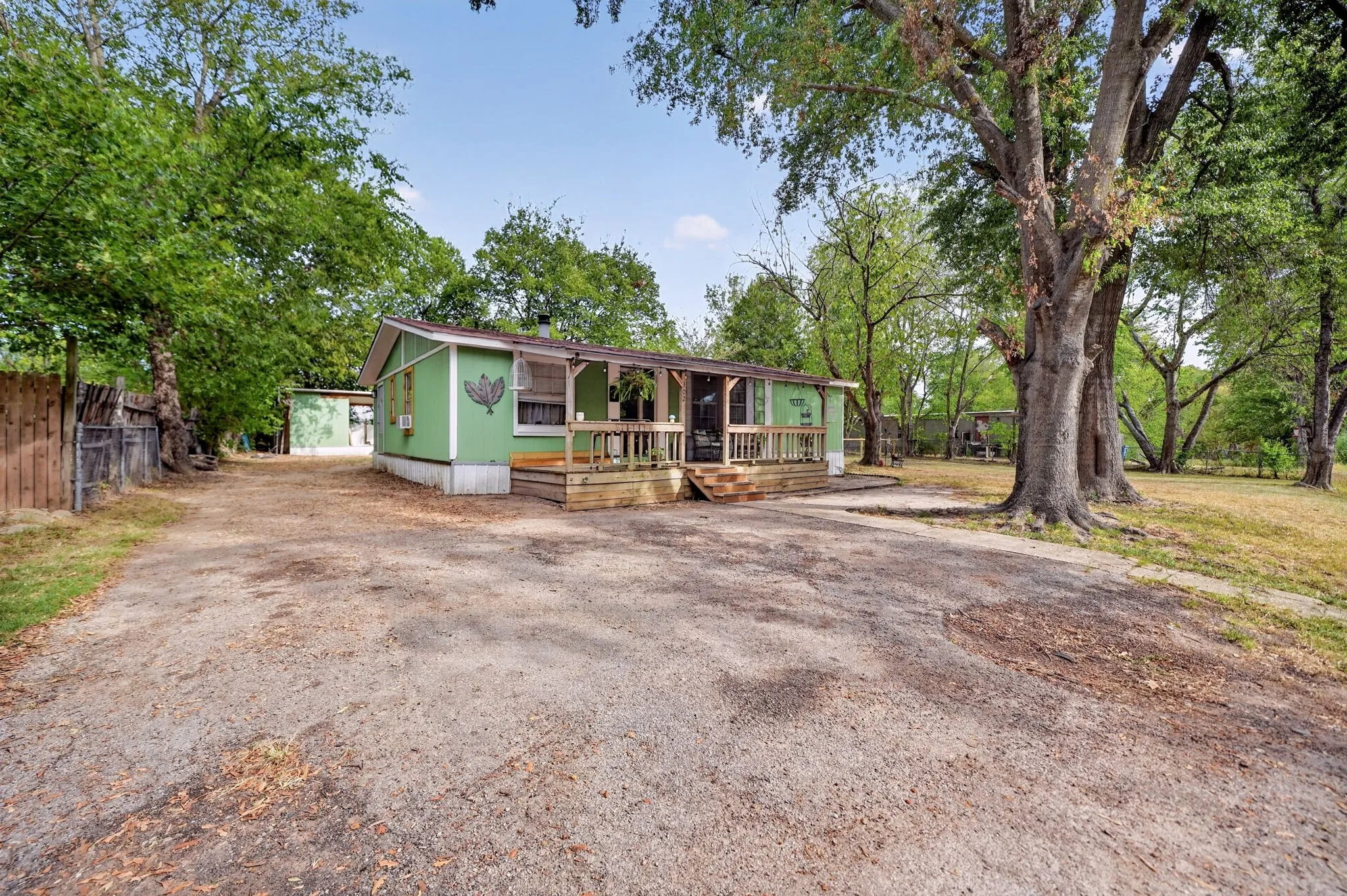 Green exterior with a covered porch, wood deck, and expansive gravel driveway