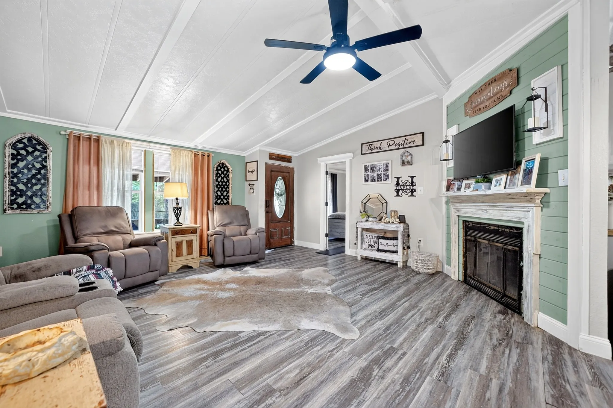 Vaulted ceilings with exposed beams, wood-finish flooring, accent wall with shiplap detail, decorative fireplace with mantel, and a built-in ceiling fan with lighting