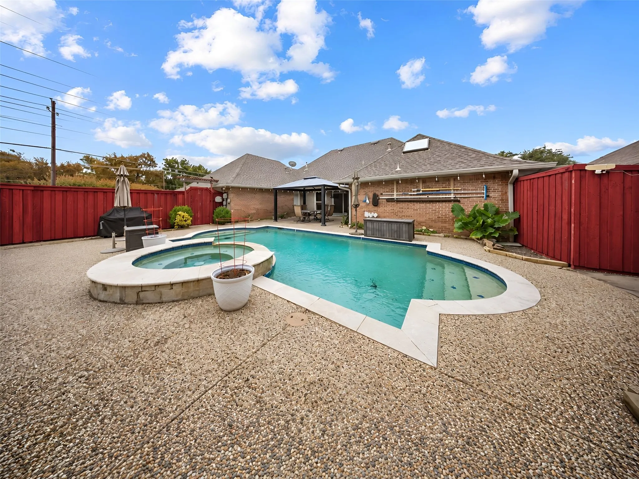 In-ground swimming pool with integrated spa, expansive pebble aggregate patio, brick exterior, and a privacy fence