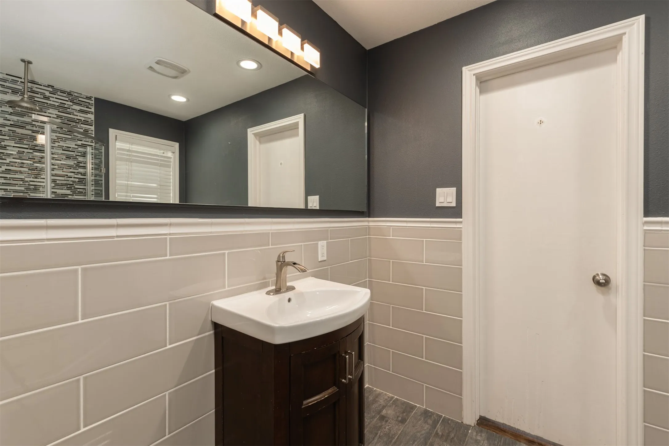 Wood-finish flooring, half-wall subway tile, dark wood vanity, white basin sink with brushed nickel faucet, and a large wall mirror