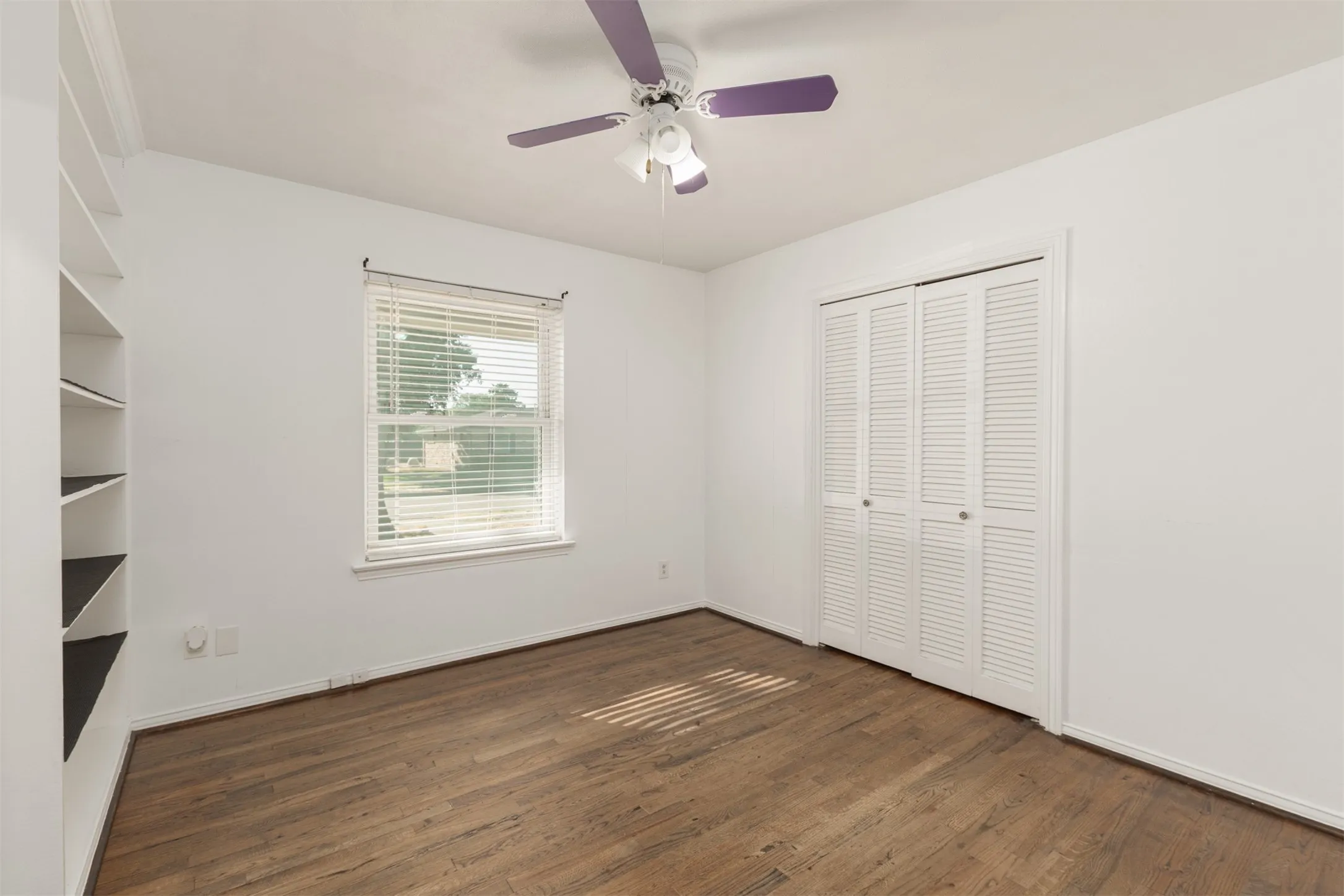 Wood-finish flooring, built-in shelving, window with blinds, bi-fold louvered closet doors, and ceiling fan