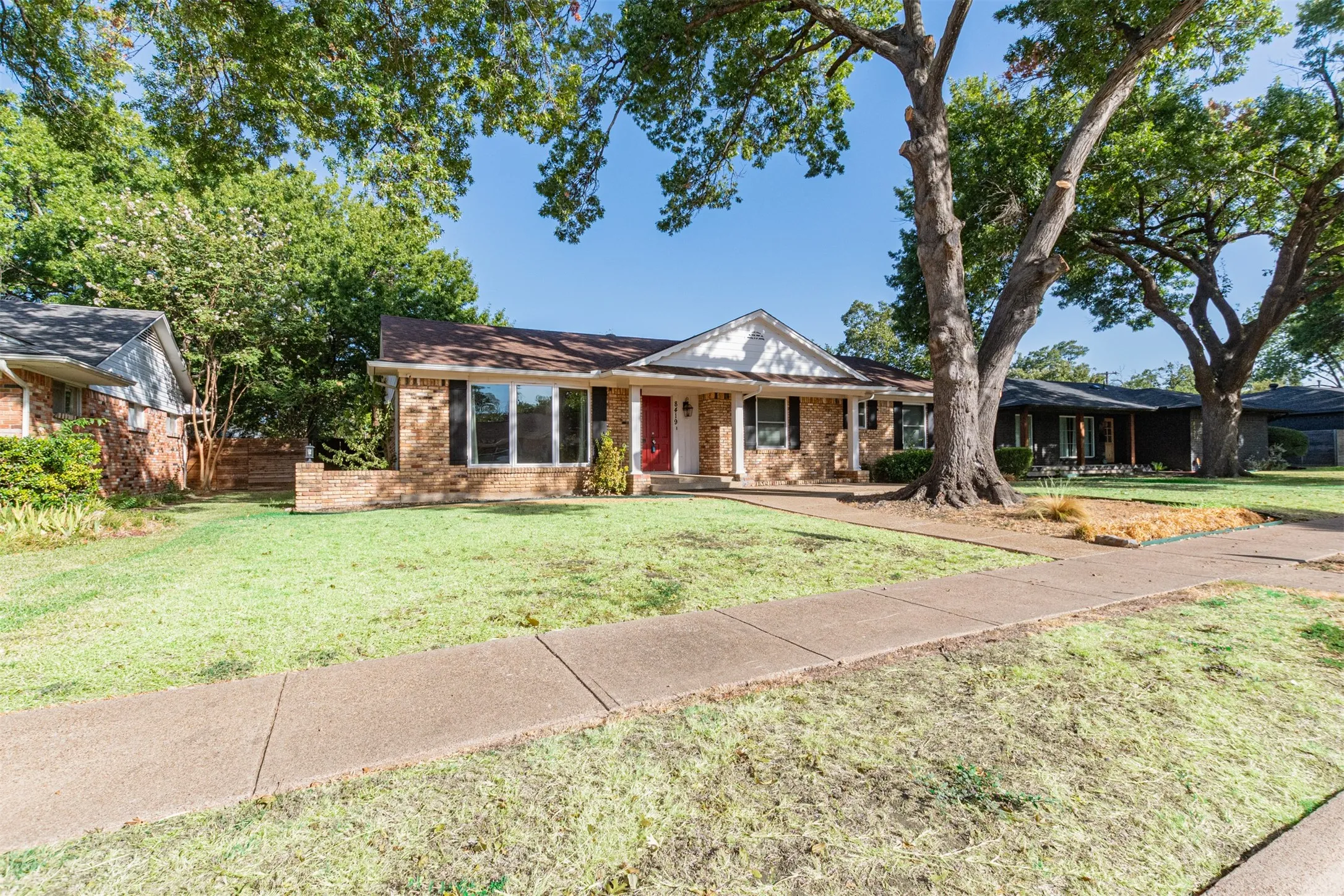 Brick facade with a red entry door, framed by mature trees, a sidewalk, and a manicured lawn