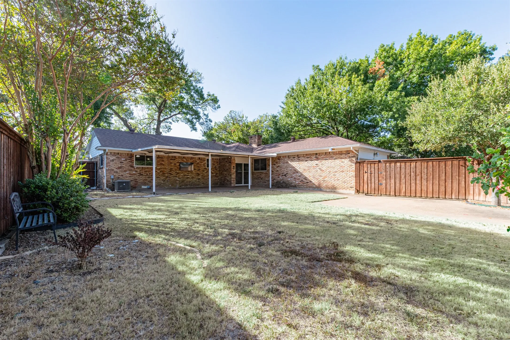 Expansive backyard featuring a covered patio, brick exterior, wood fencing, and mature trees