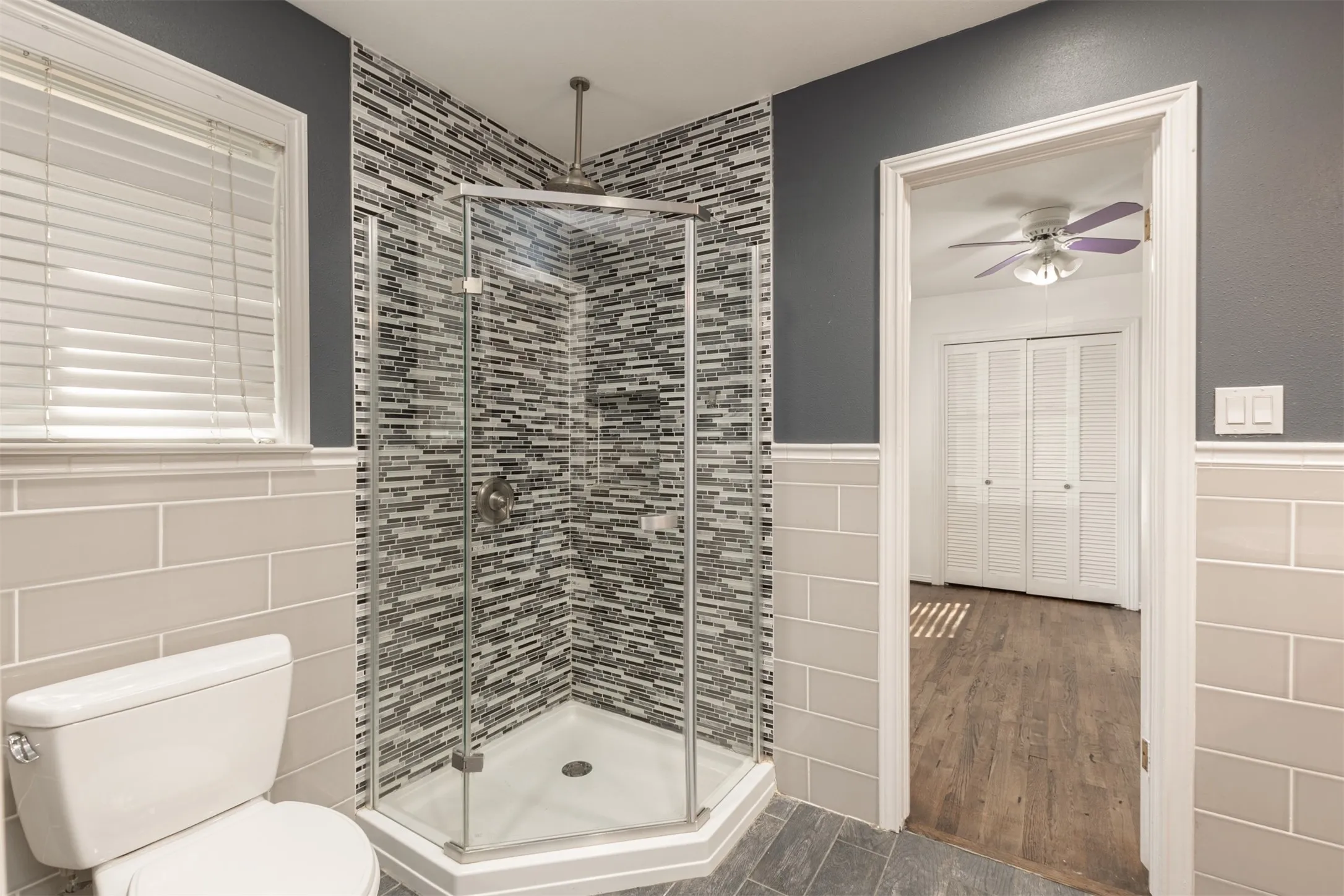 Corner glass-enclosed shower with mosaic tile surround, white toilet, and light gray subway tile wainscoting