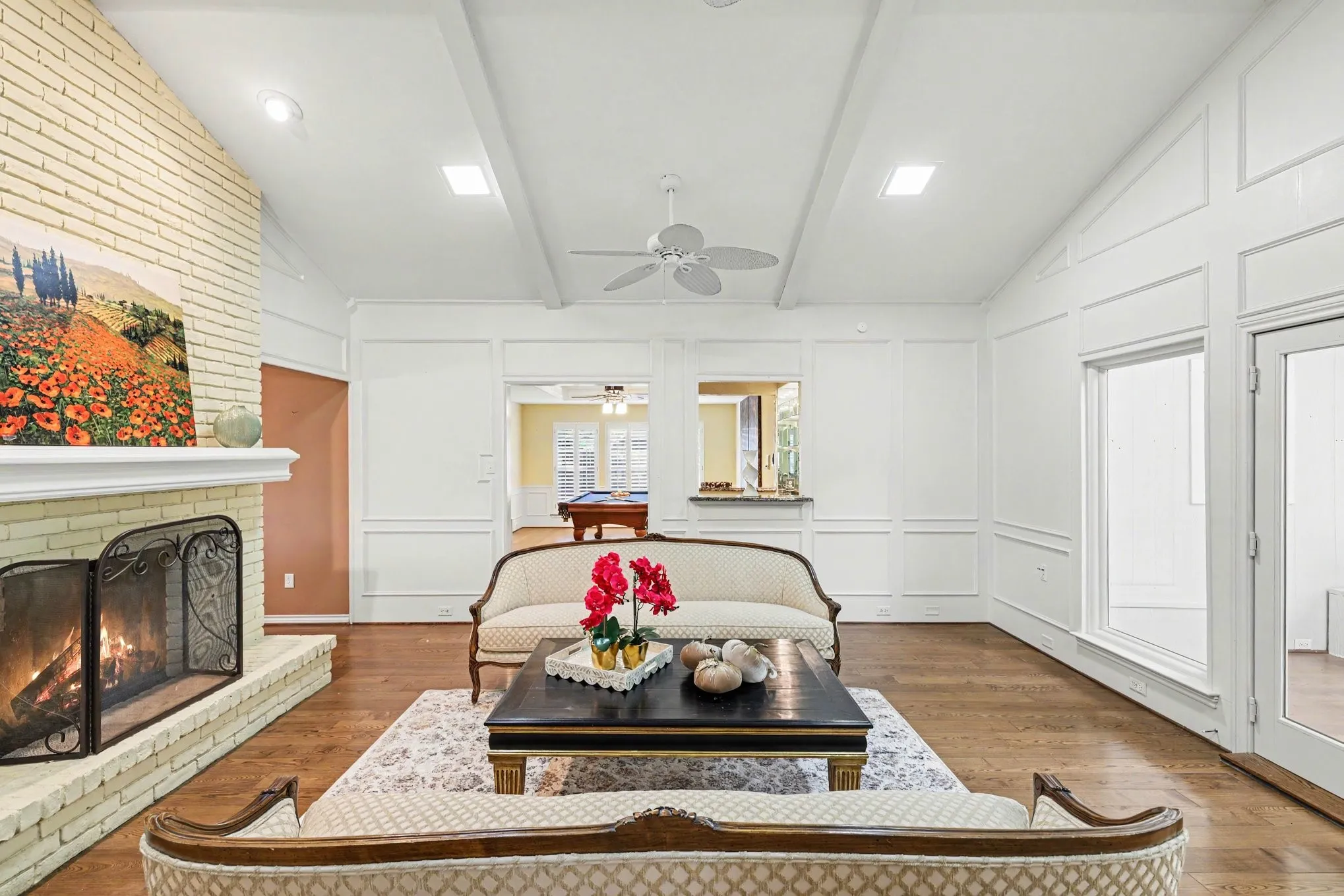 Vaulted ceiling with exposed beams, brick fireplace, wood-finish flooring, recessed lighting, and white wall paneling