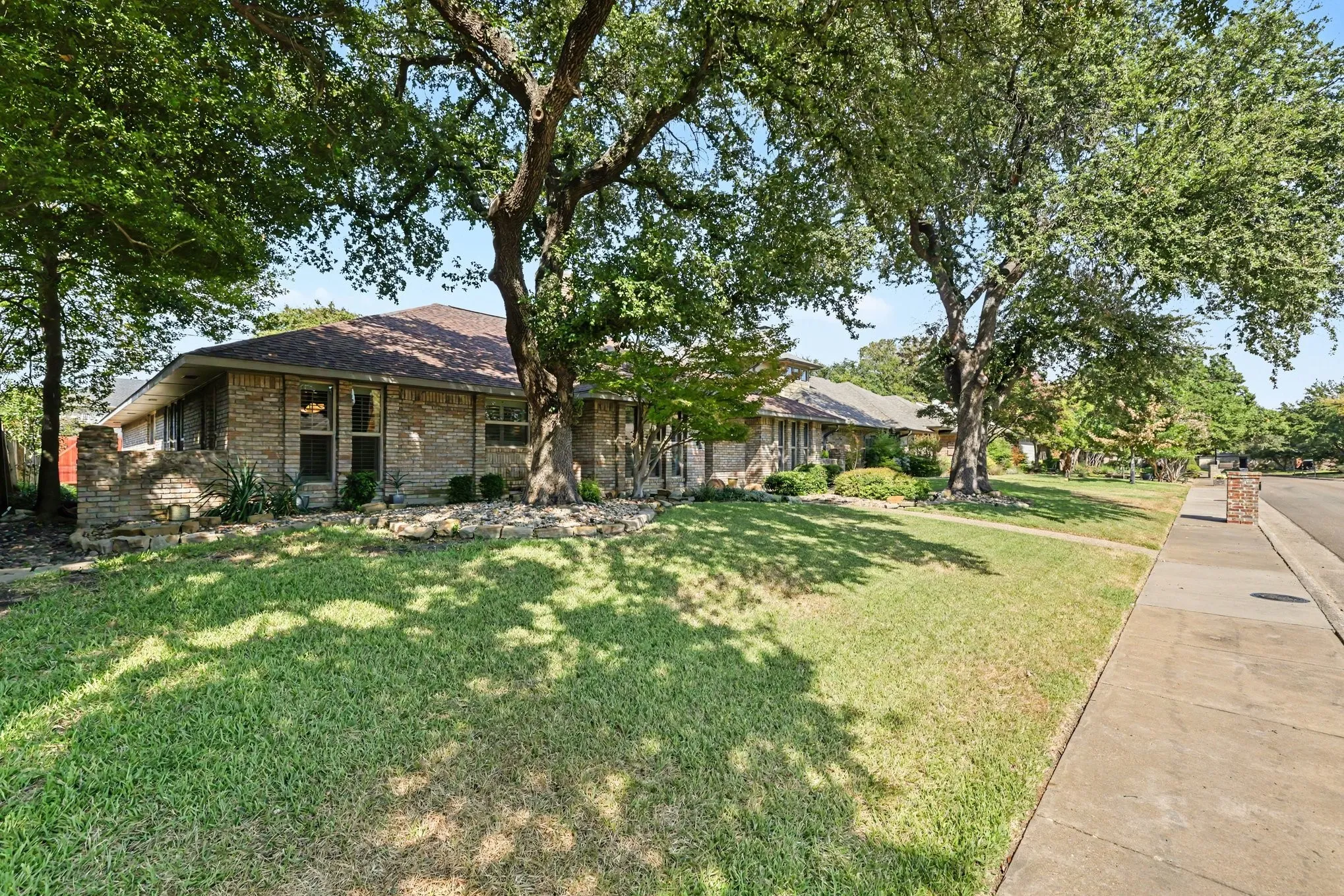 Brick exterior with a shingle roof, mature trees, and a concrete sidewalk