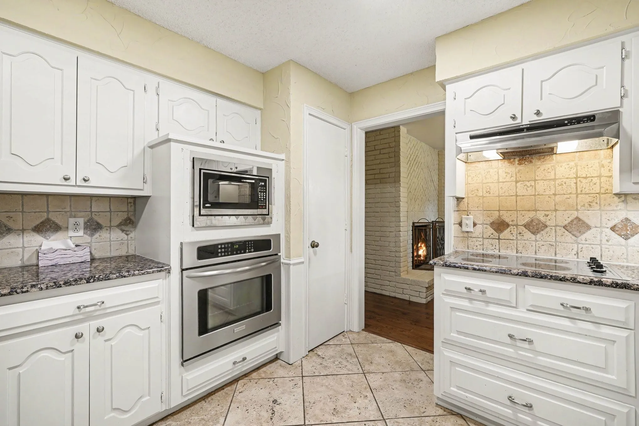 White cabinetry with decorative fronts, stainless steel built-in appliances, tiled flooring, speckled countertops, and a tiled backsplash