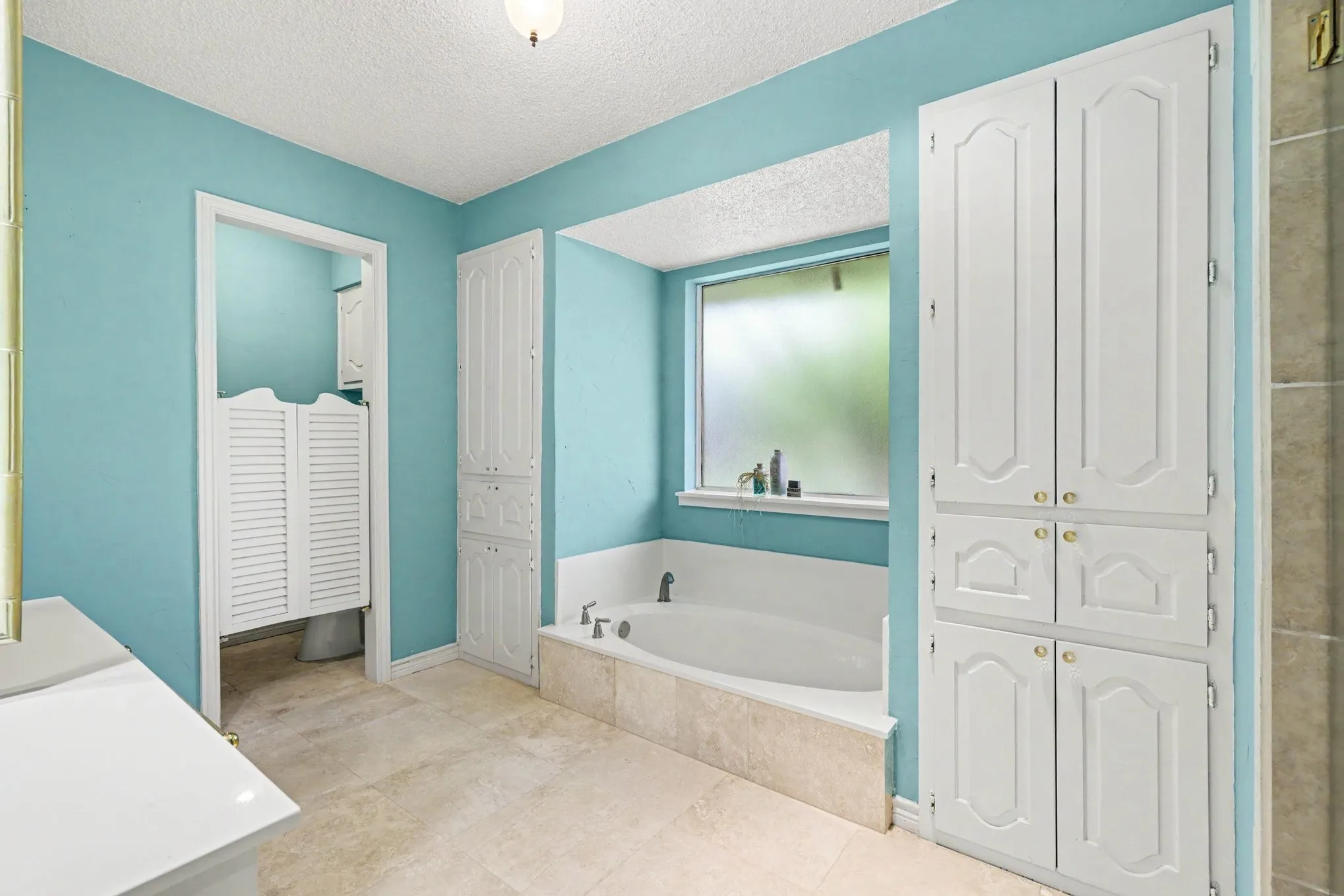 Built-in soaking tub with surrounding tile, large obscured window, multiple built-in storage cabinets, and tile flooring