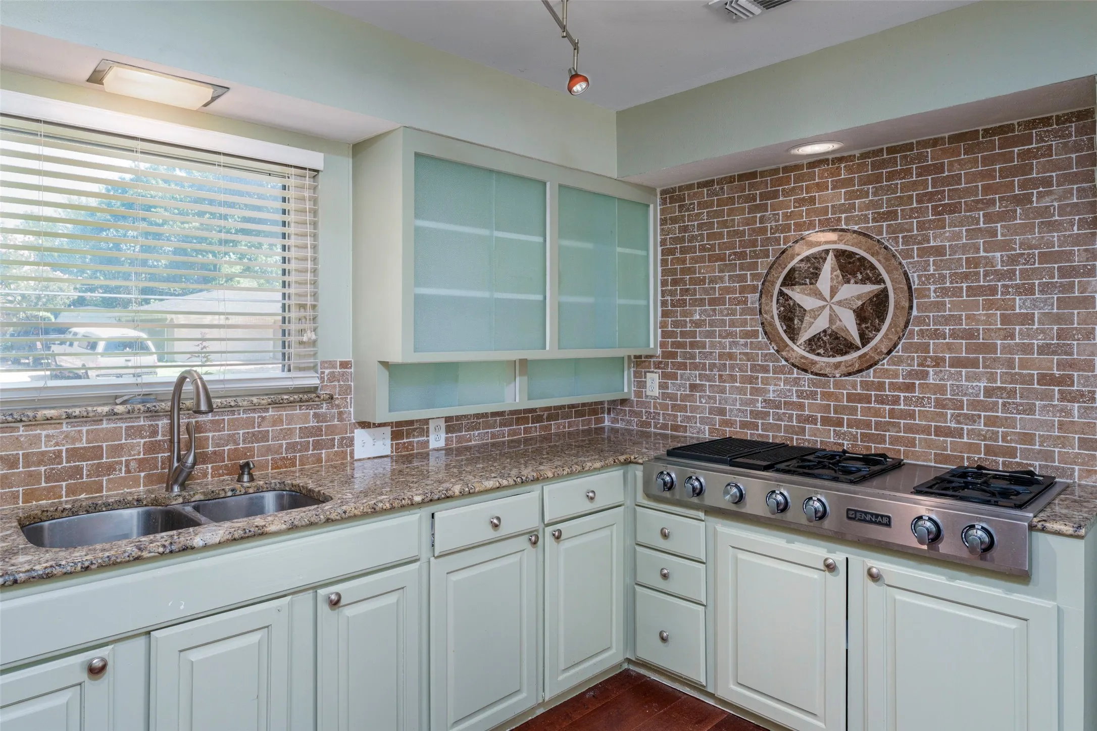 Kitchen featuring light-toned cabinetry, a stainless steel gas range, a dual basin stainless steel sink, granite countertops, and a brick-patterned backsplash with a decorative star inlay