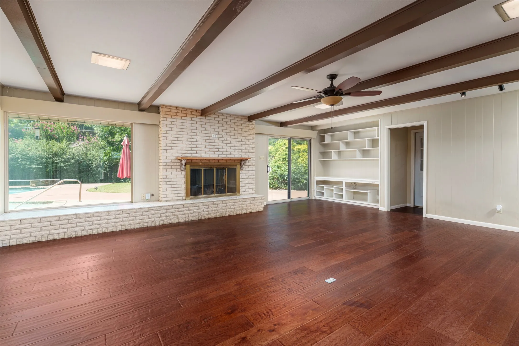 Exposed beam ceiling, wood-finish flooring, brick fireplace with a wood mantle, built-in shelving, and large windows