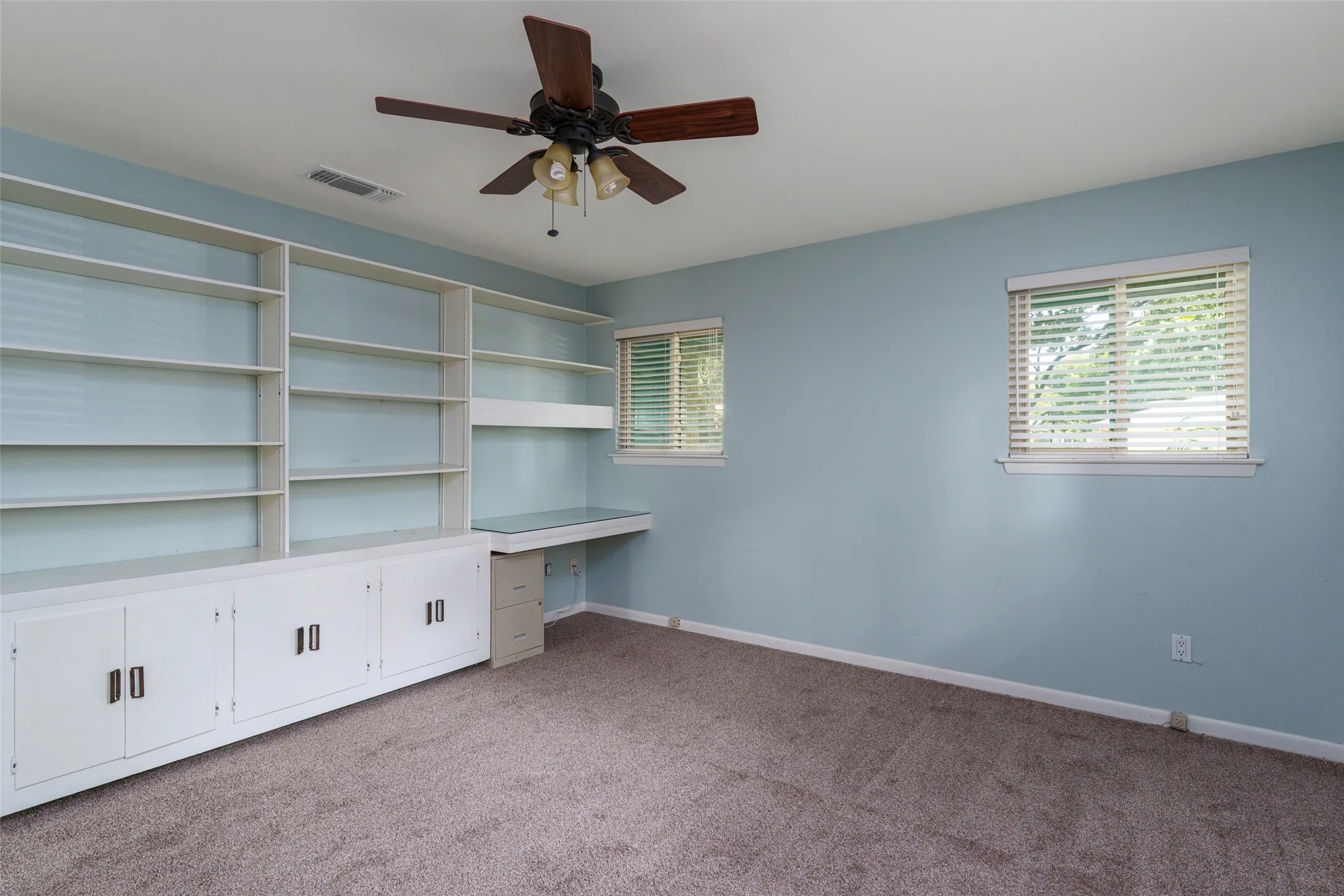 Built-in shelving and cabinetry, integrated desk space, ceiling fan, and two windows with blinds