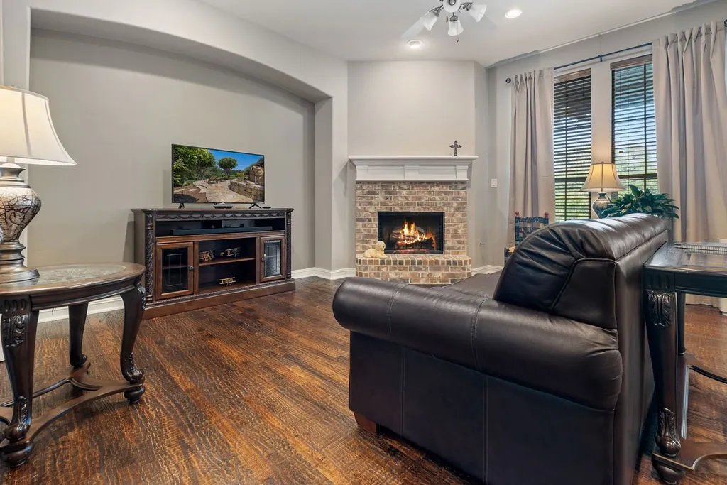 Wood-finish flooring, brick fireplace with a white mantel, ceiling fan, and large windows with blinds