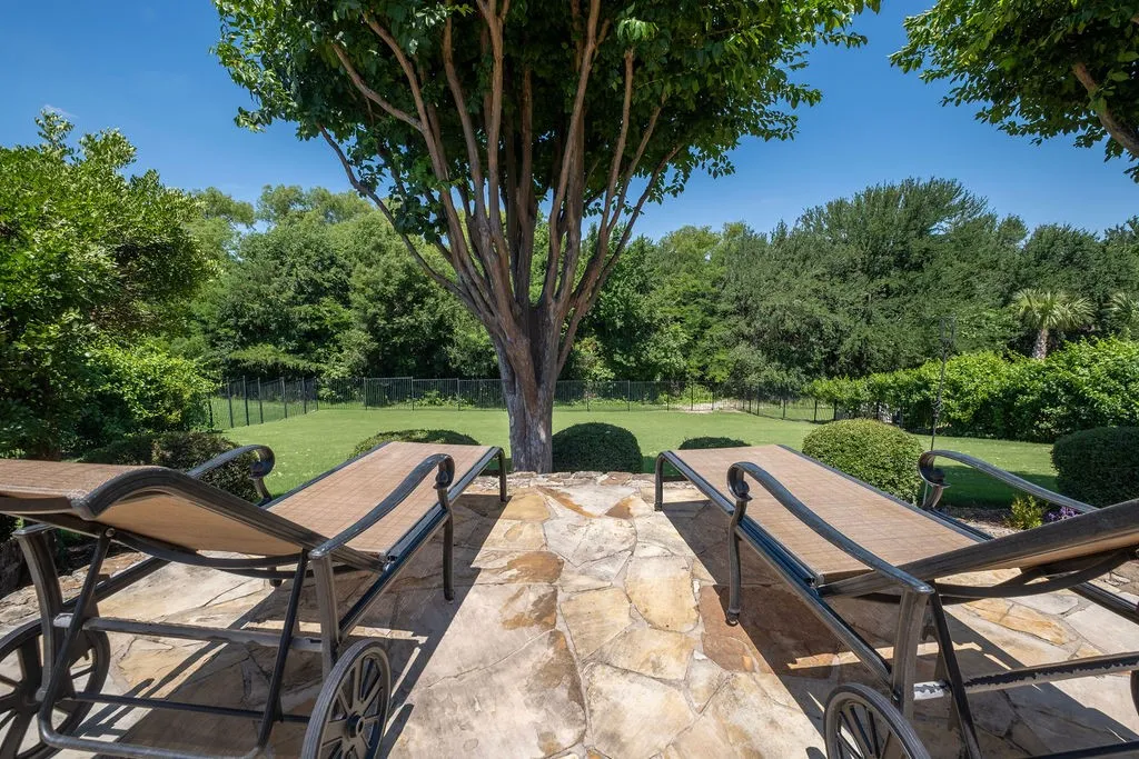 Expansive outdoor hardscaping with natural stone paving, mature trees, manicured lawn, and a perimeter fence