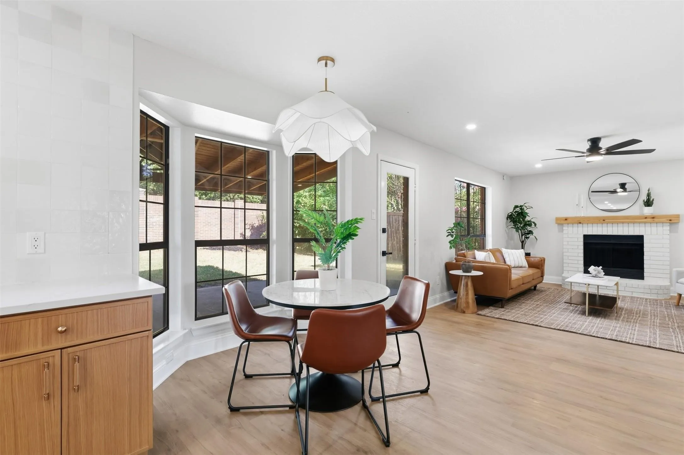 Open-concept living area featuring light wood-finish flooring, a white brick fireplace with a wood mantle, and a ceiling fan