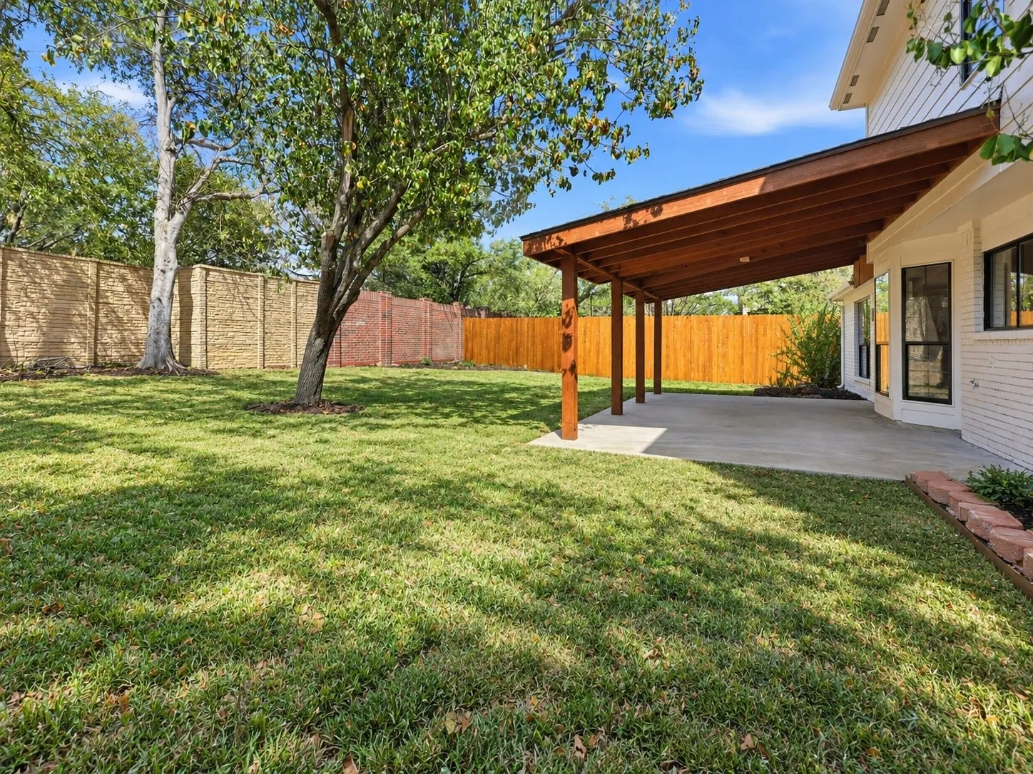 Expansive backyard featuring a concrete patio with a wood-beam covered structure, mature trees, and a brick-patterned retaining wall