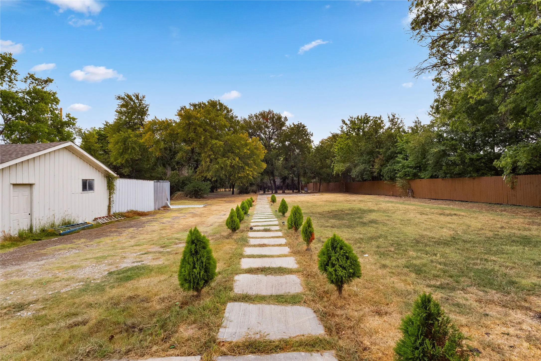 Expansive outdoor area featuring a stepping stone pathway, mature trees, a privacy fence, and an accessory building