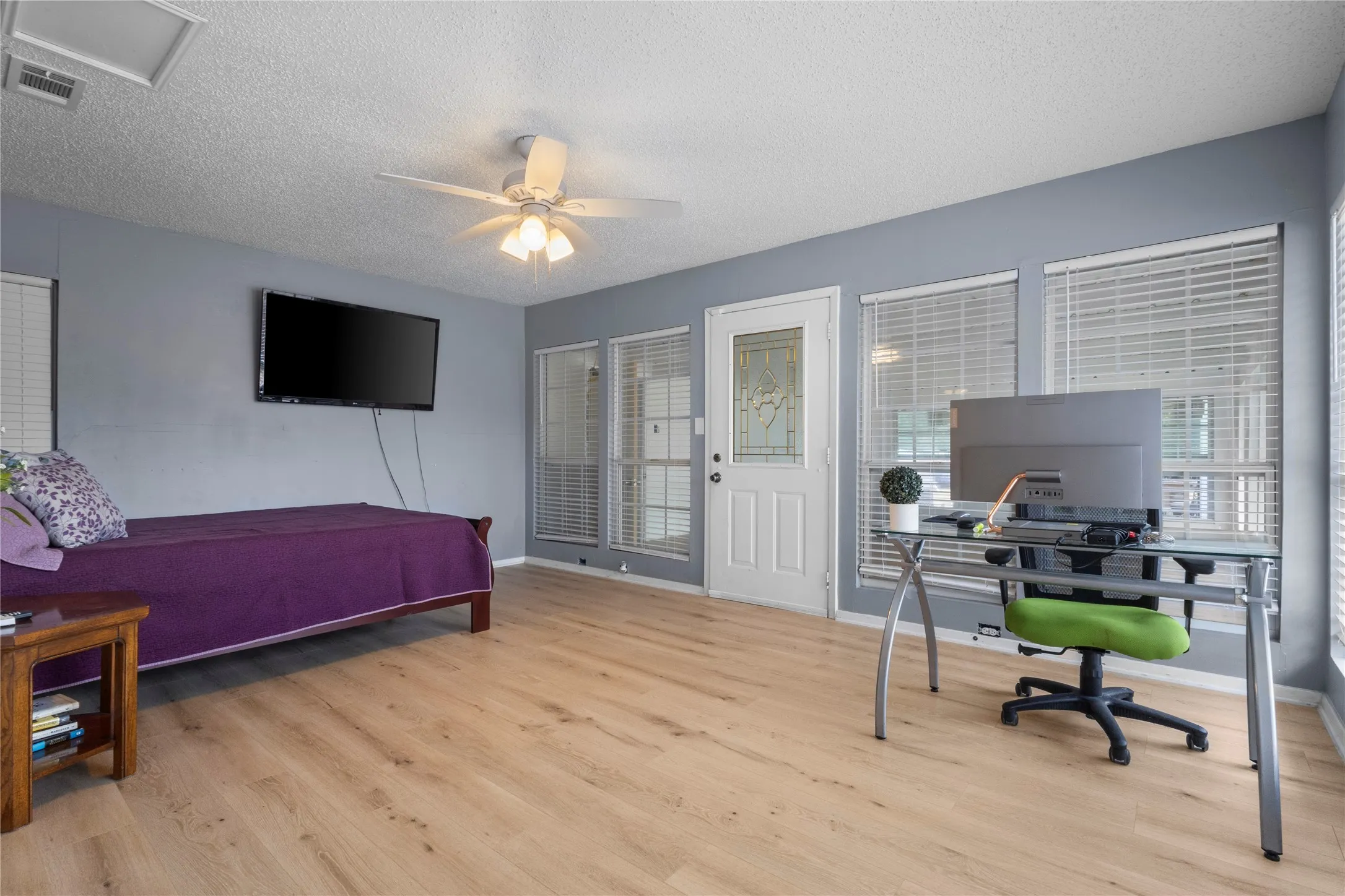 Wood-finish flooring throughout, ceiling fan with integrated lighting, multiple windows with blinds, and a white paneled door with decorative glass