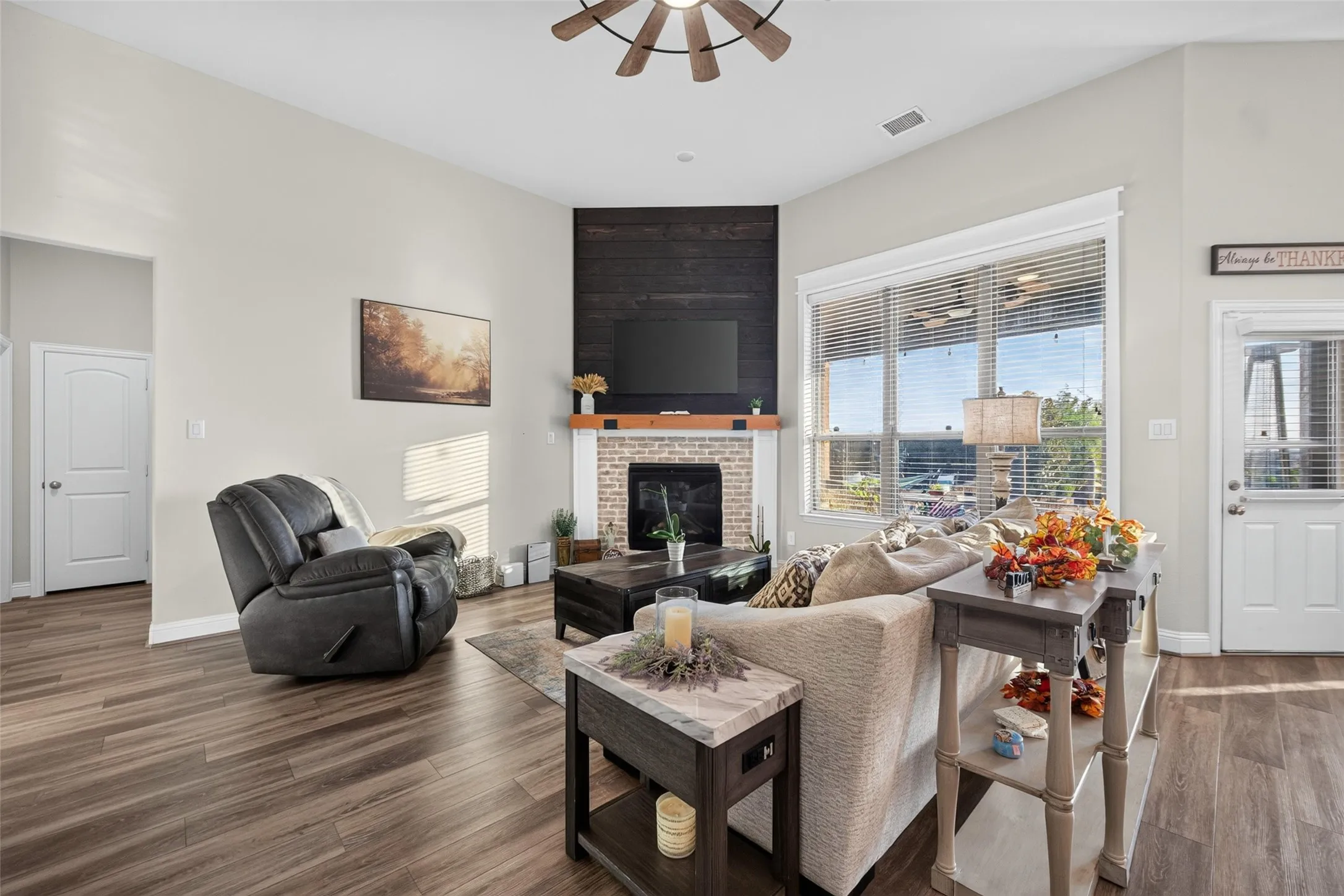 Vaulted ceiling with a ceiling fan, corner fireplace with a wood-finish mantel and brick surround, large windows with horizontal blinds, and wood-finish flooring