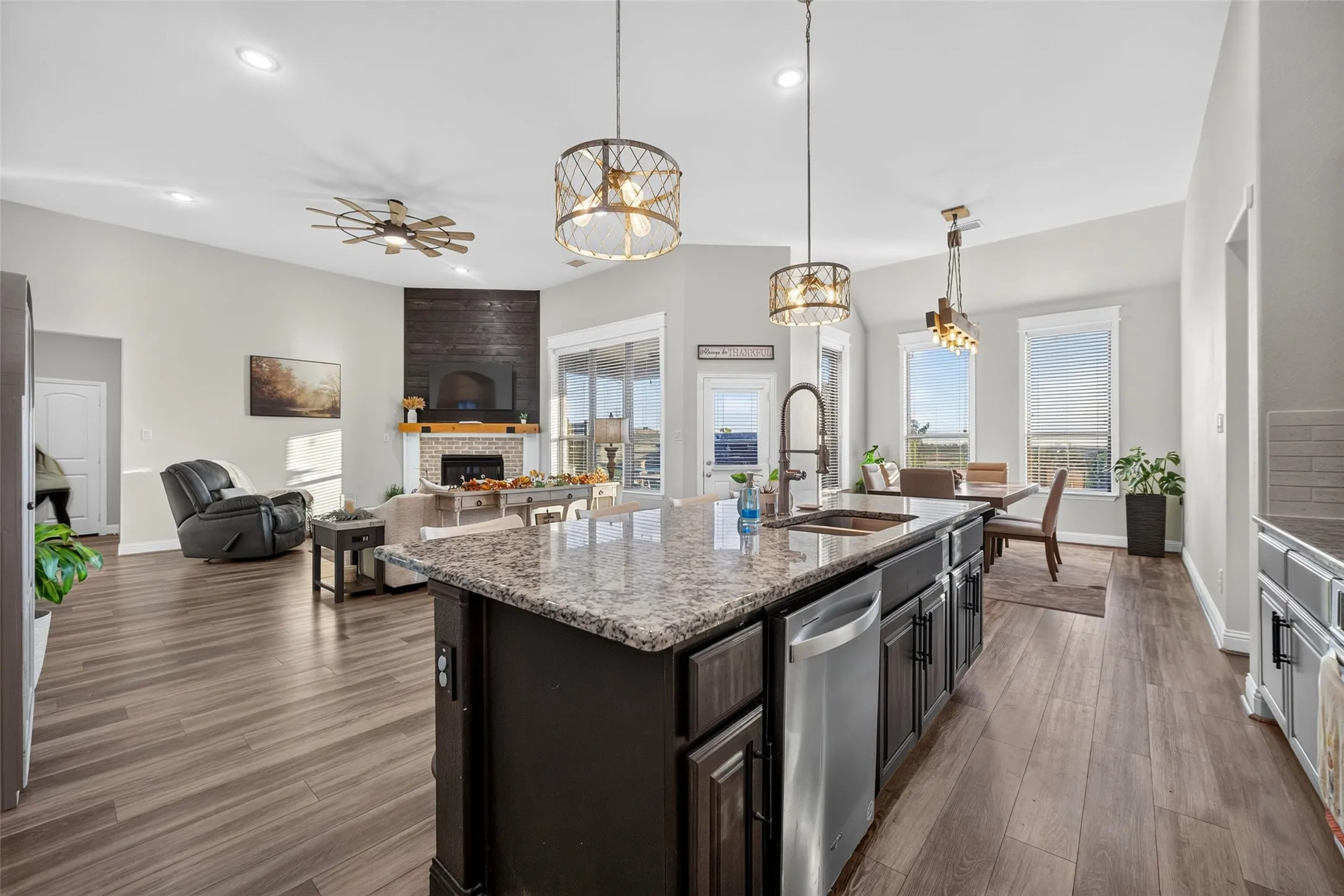 Open-concept kitchen and living area featuring a large island with stone countertop and stainless steel appliances, wood-finish flooring throughout, pendant lighting, and expansive windows with natural light