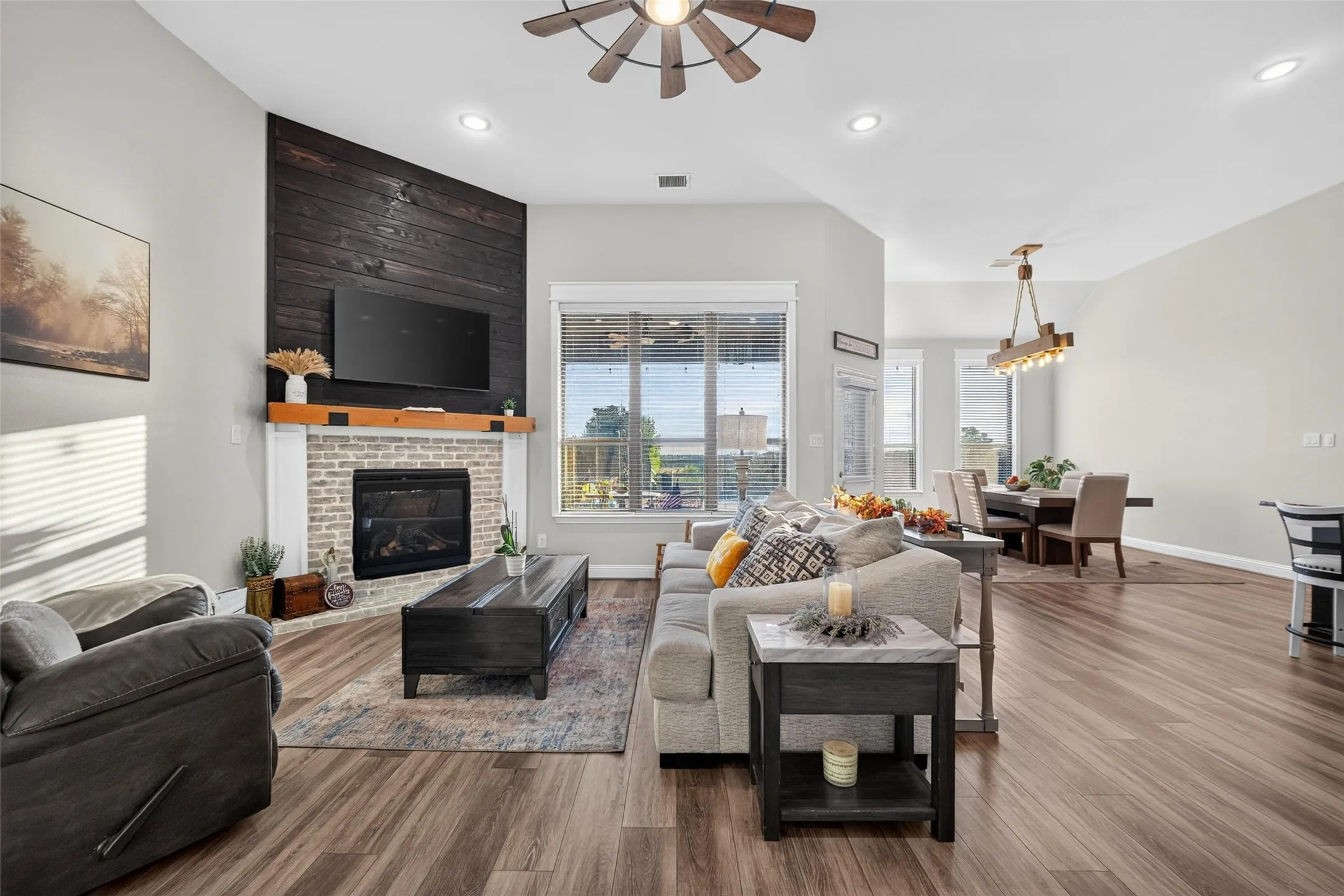 Vaulted ceilings with recessed lighting, wood-finish flooring, brick fireplace with a wood mantle, shiplap accent wall, and a ceiling fan