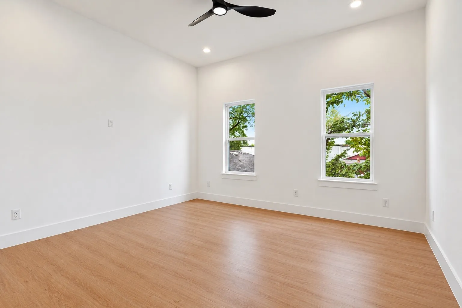 Light wood-finish flooring, two large windows, white walls, recessed lighting, and a ceiling fan