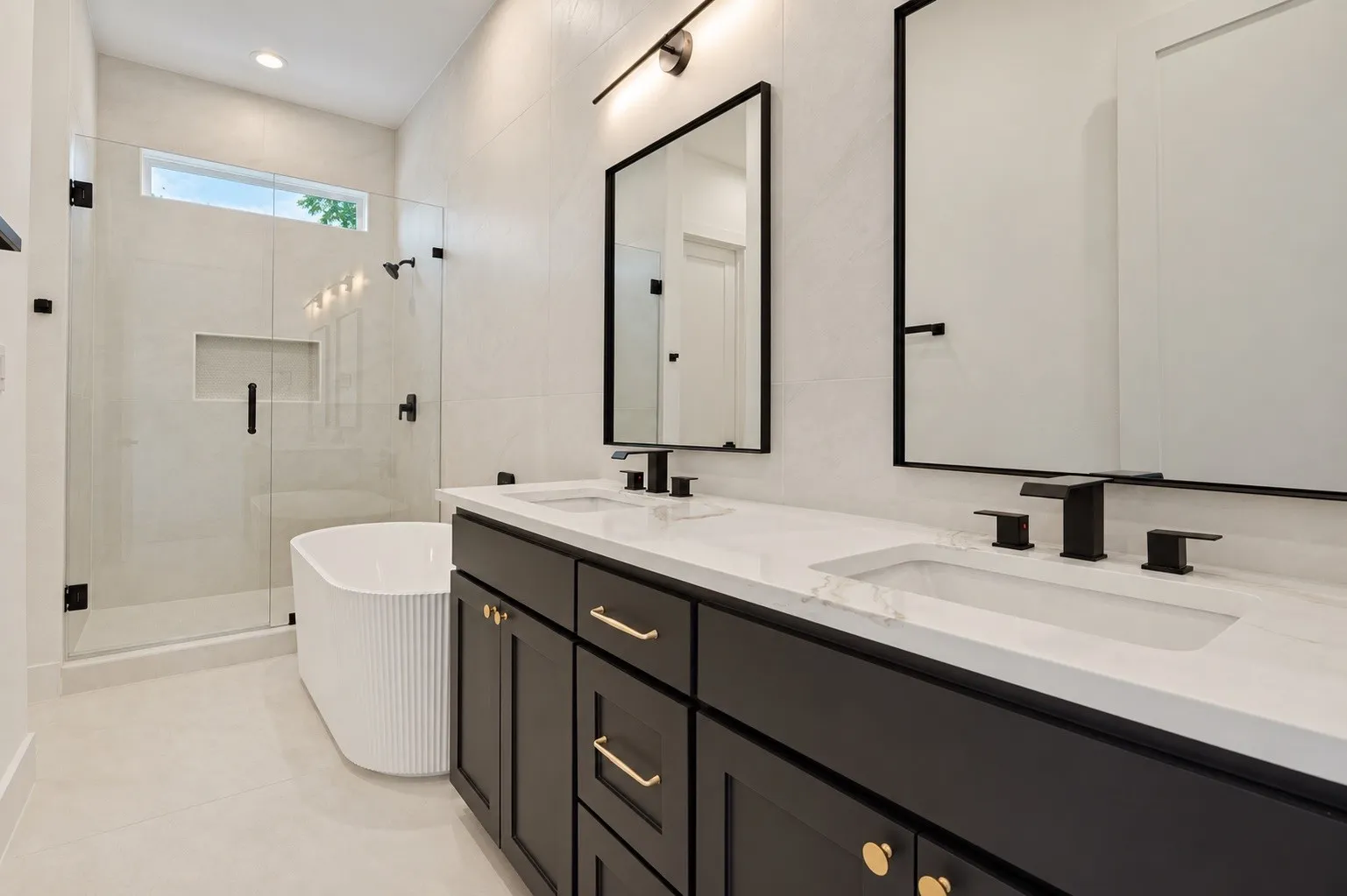 Dual vanity with integrated sinks and black cabinetry, frameless shower with glass enclosure, freestanding fluted bathtub, transom window, and tiled walls