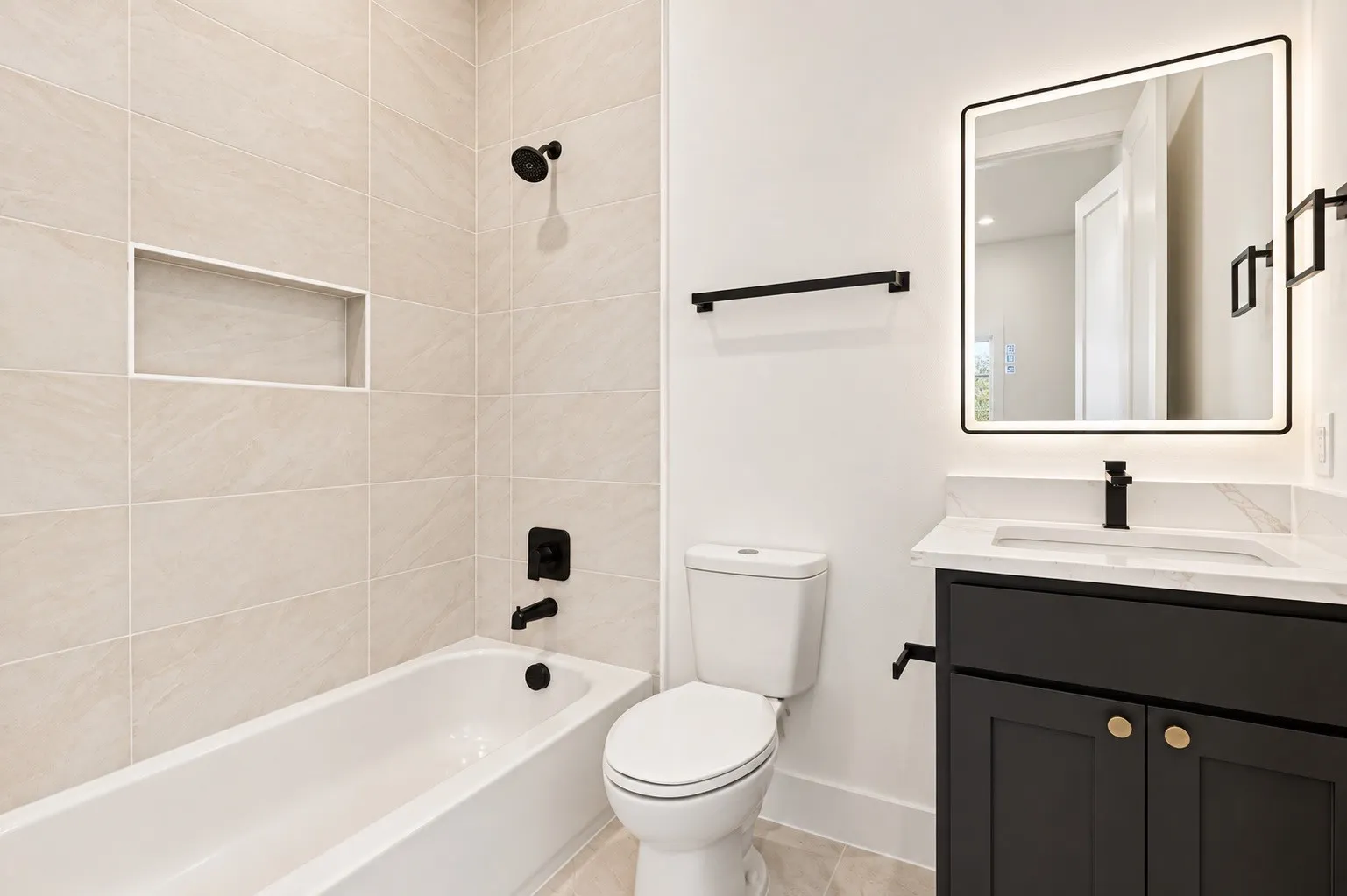 Bathroom featuring a tiled shower surround with a built-in niche, white bathtub, black vanity with a white countertop, and an illuminated mirror