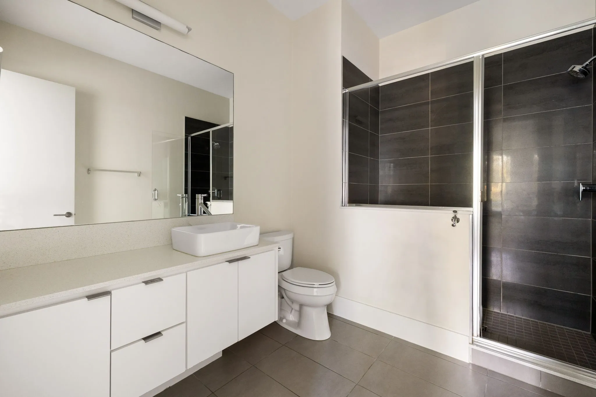 Rectangular vessel sink, white cabinetry with silver-tone hardware, large vanity mirror, tile flooring, and a glass-enclosed shower with dark-tiled walls