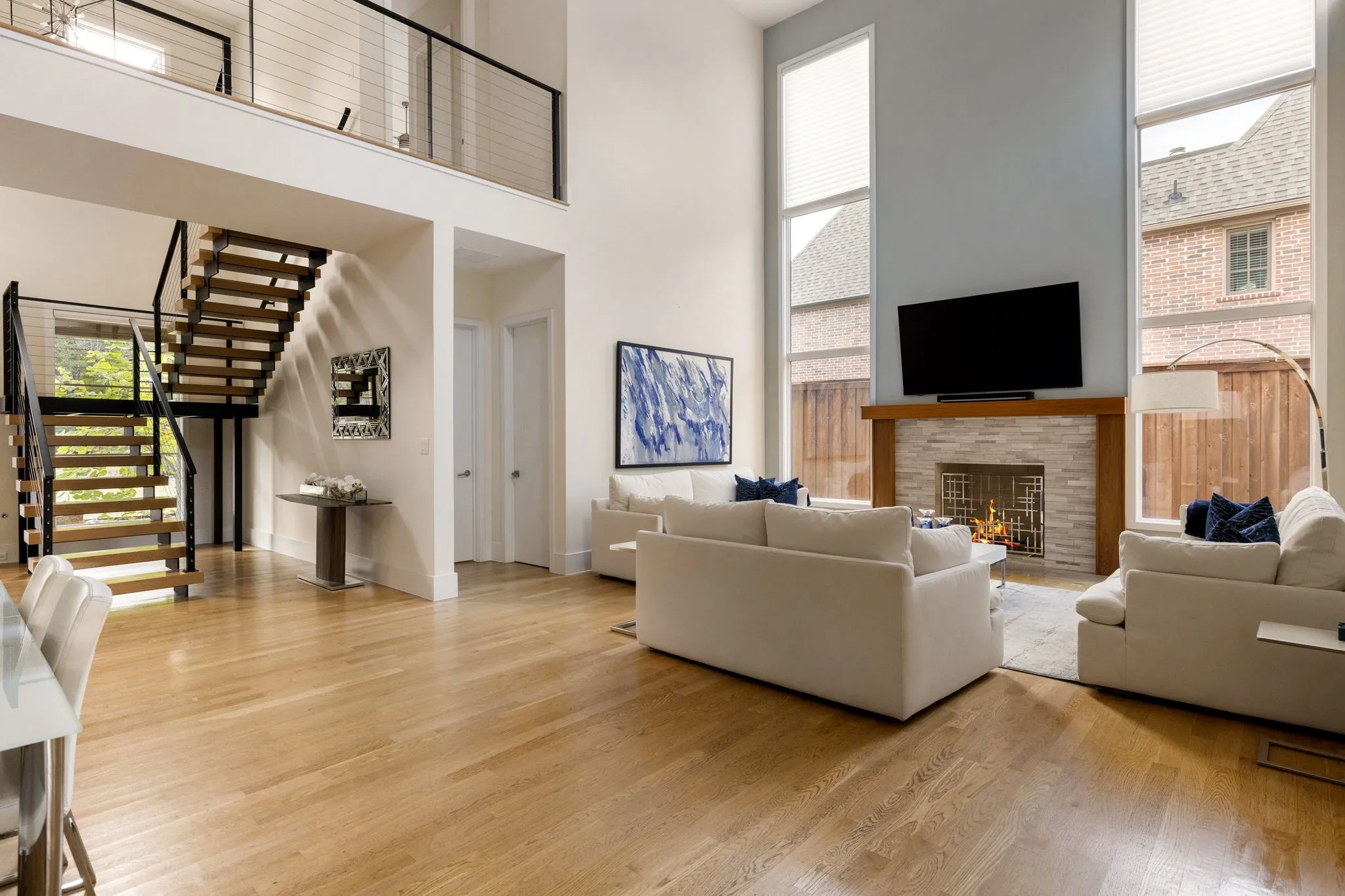 Open-concept living area featuring high ceilings, wood-finish flooring, a contemporary floating staircase with cable railing, a stone-clad fireplace, and tall windows
