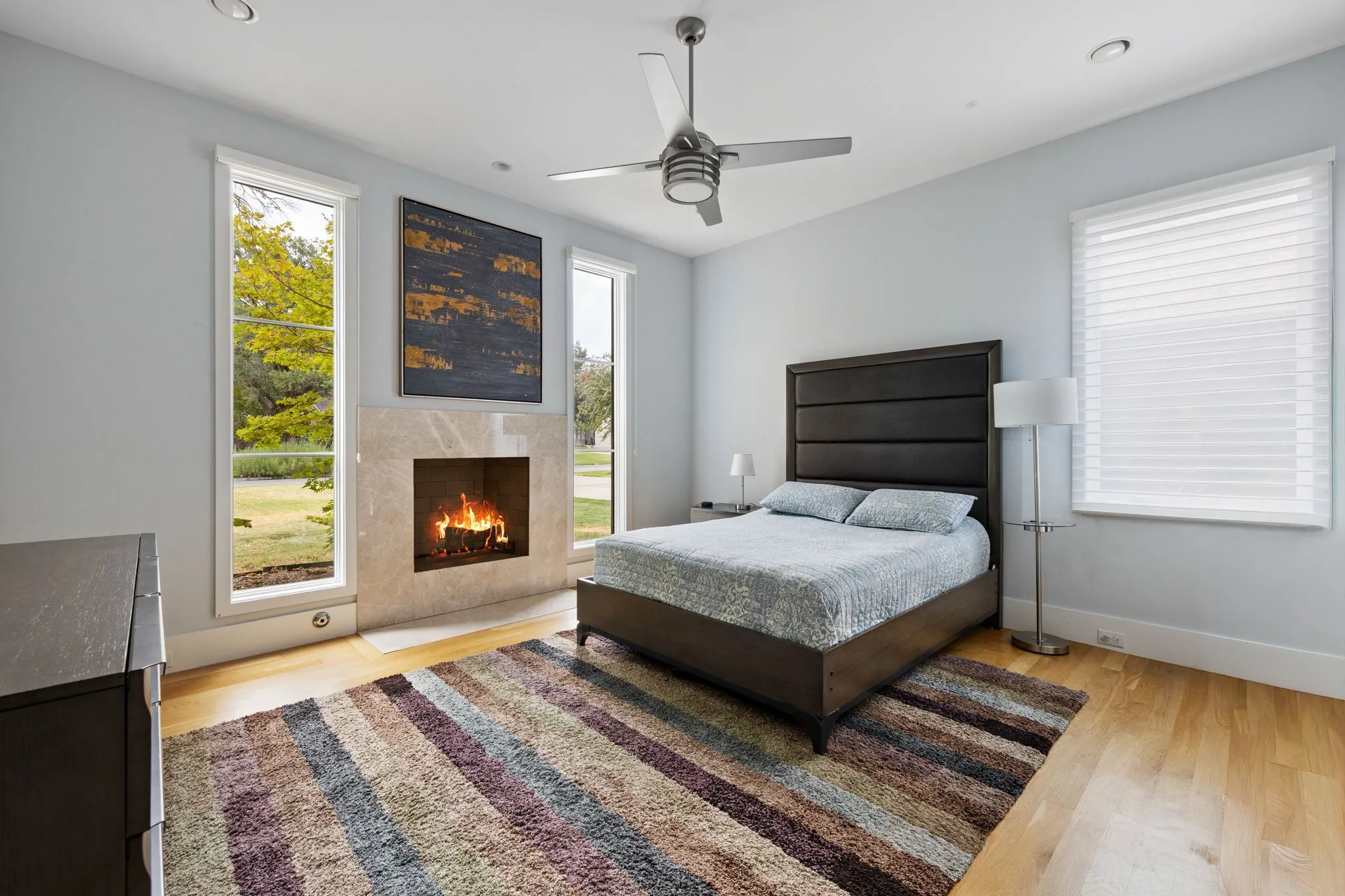 Primary bedroom featuring a wood-finish floor, gas fireplace with stone surround, ceiling fan with integrated lighting, and abundant natural light from dual windows with motorized shades