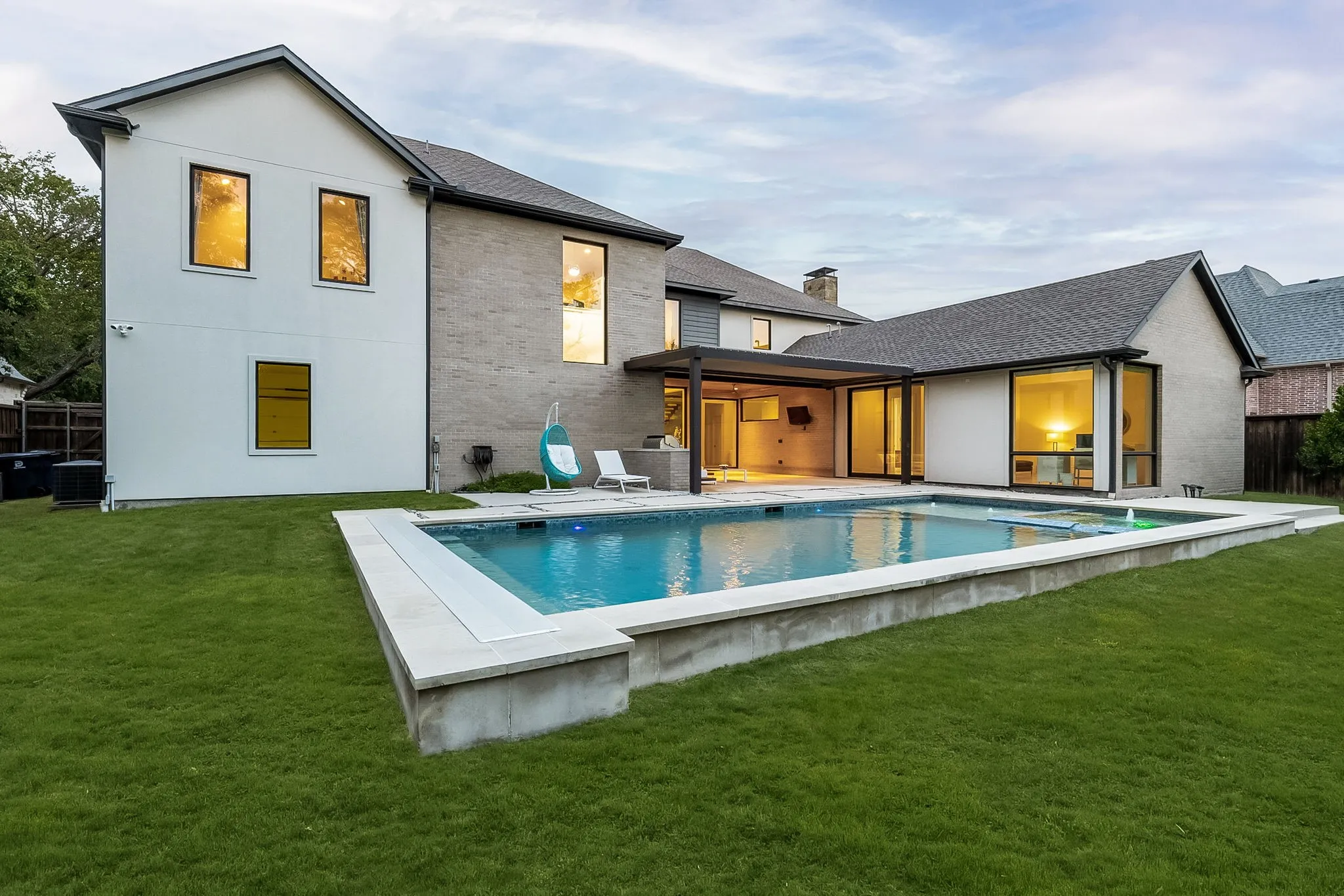 Expansive swimming pool with a surrounding concrete and stone border, large grass area, covered patio, and extensive glass doors